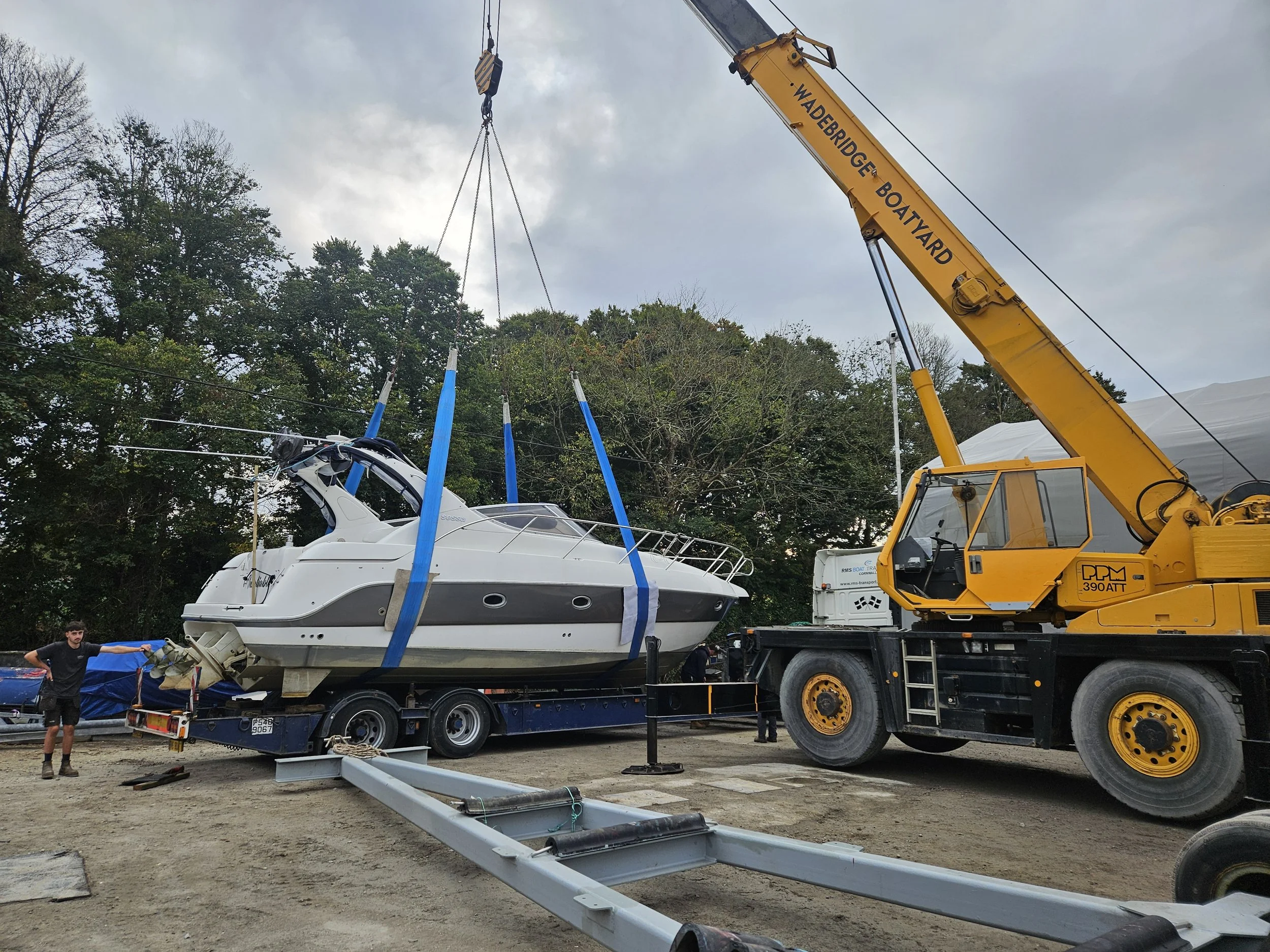 A large yellow crane lifting a white yacht onto a flatbed trailer at a boatyard with trees in the background.