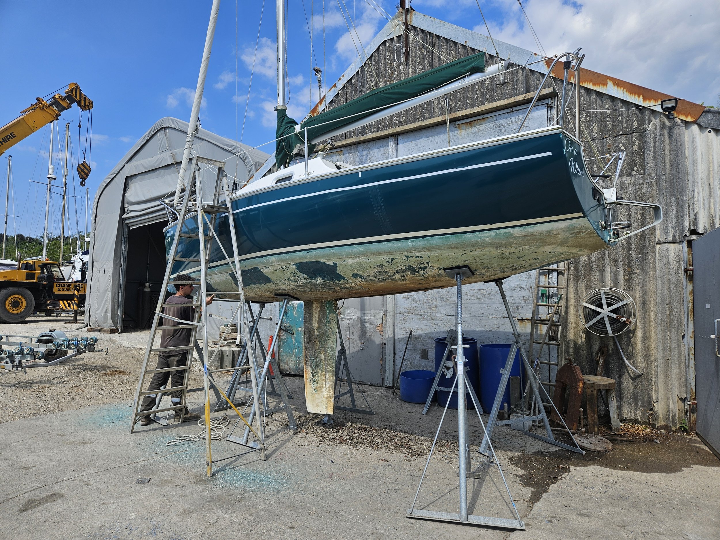 A sailboat on supports outside a boat repair shop, surrounded by tools and equipment, with a person working nearby.