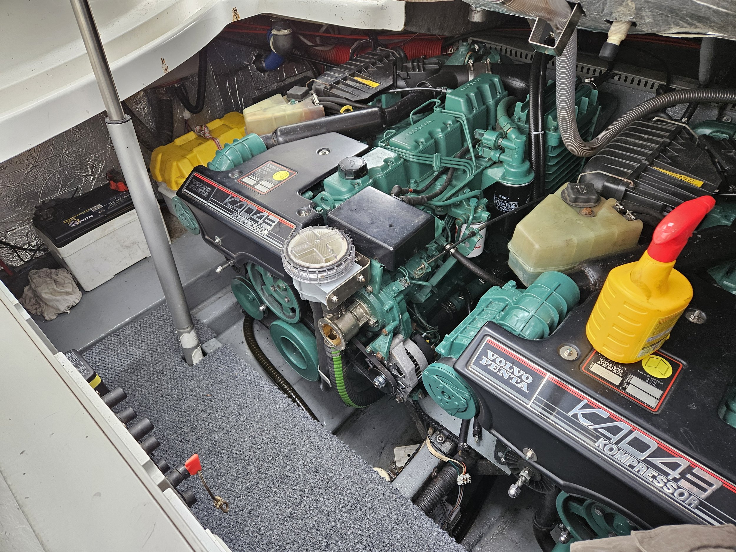 Marine engine room showing two green engines labeled V8 KAD 43 Volvo Penta, with various hoses, a yellow oil container, battery, and other mechanical components.