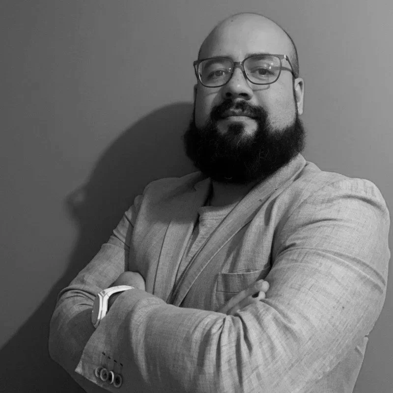 A man with glasses and a beard wearing a light-colored blazer, standing with arms crossed against a plain wall.