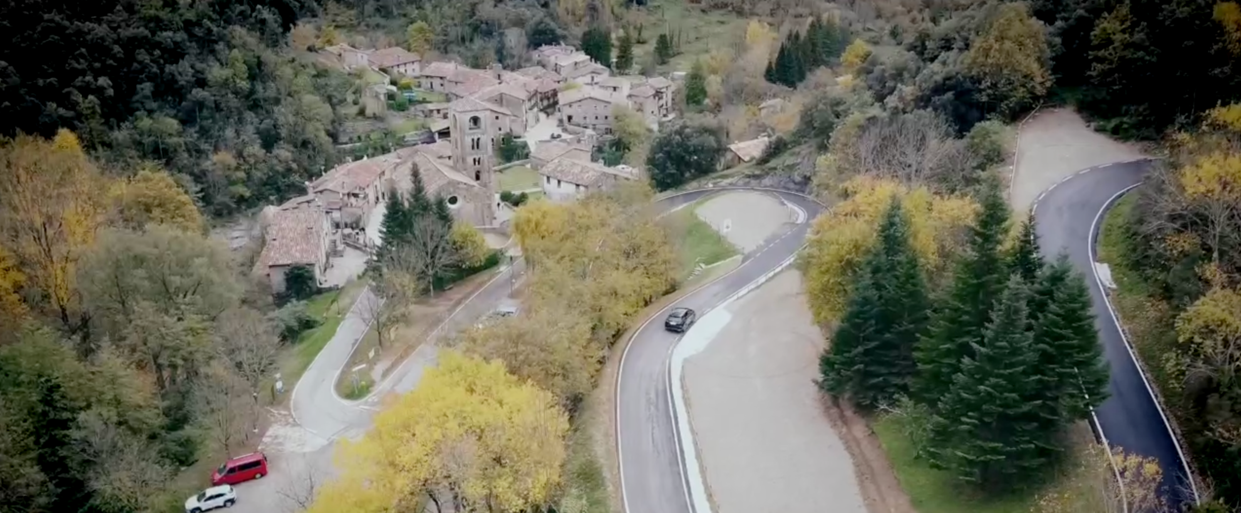 Vista aérea de un pueblo en una zona montañosa con árboles en colores de otoño y carreteras serpenteantes.