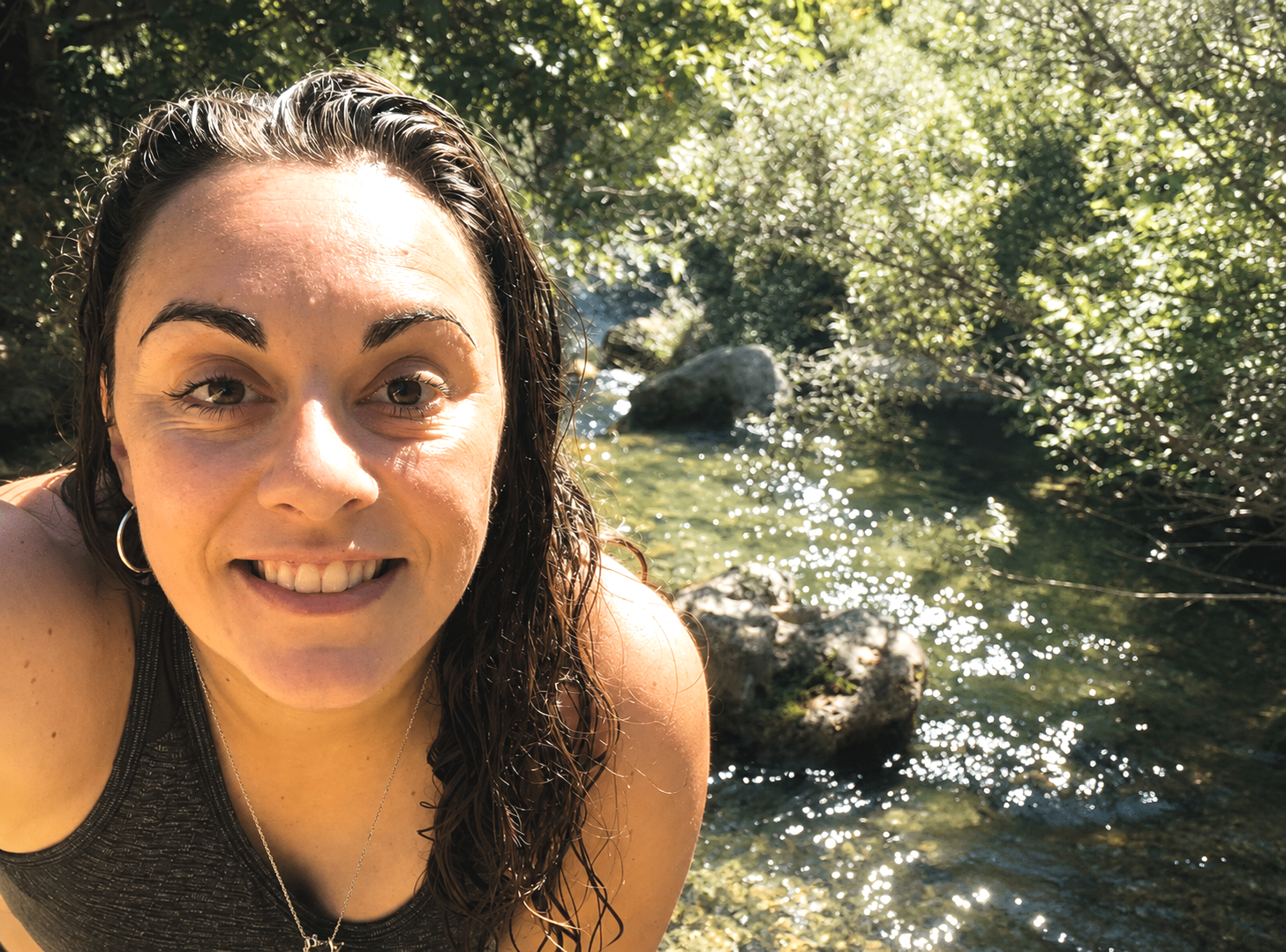 Una mujer con cabello húmedo en un entorno natural cerca de un río, con árboles y agua brillante en el fondo.