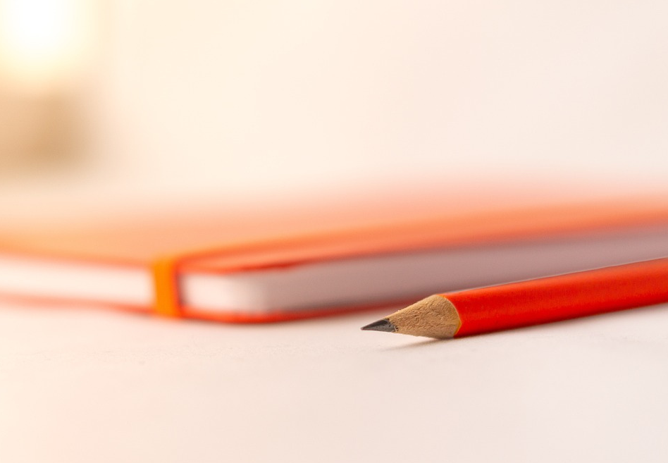 Un crayon posé à coté d'un carnet orange. Prendre le temps de l'analyse, de la reflexion avant de proposer des solutions.