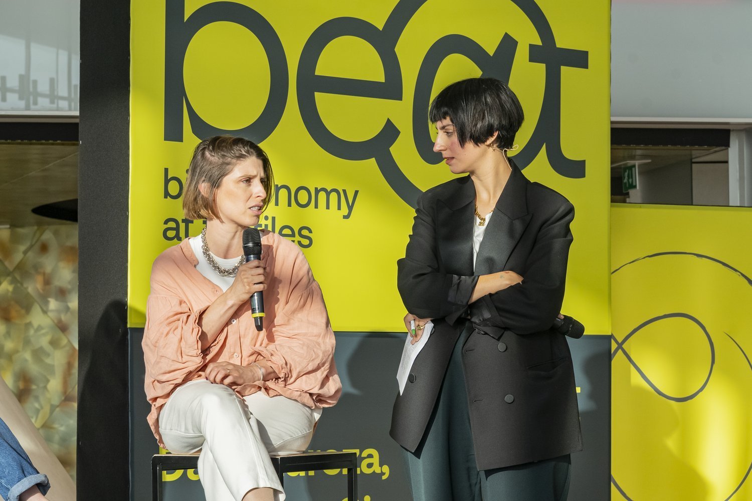 Duas mulheres em uma discussão na frente de um fundo amarelo com texto sobre economia. Uma está sentada, segurando um microfone, e a outra está de pé com os braços cruzados.