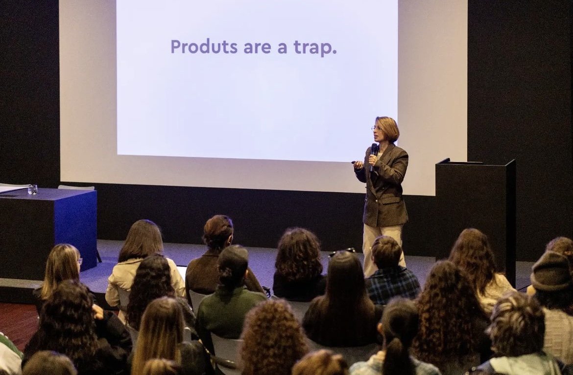 Mulher apresentando uma palestra diante de uma tela que diz 'Produtos são uma armadilha', para uma audiência sentada em uma sala de conferências.