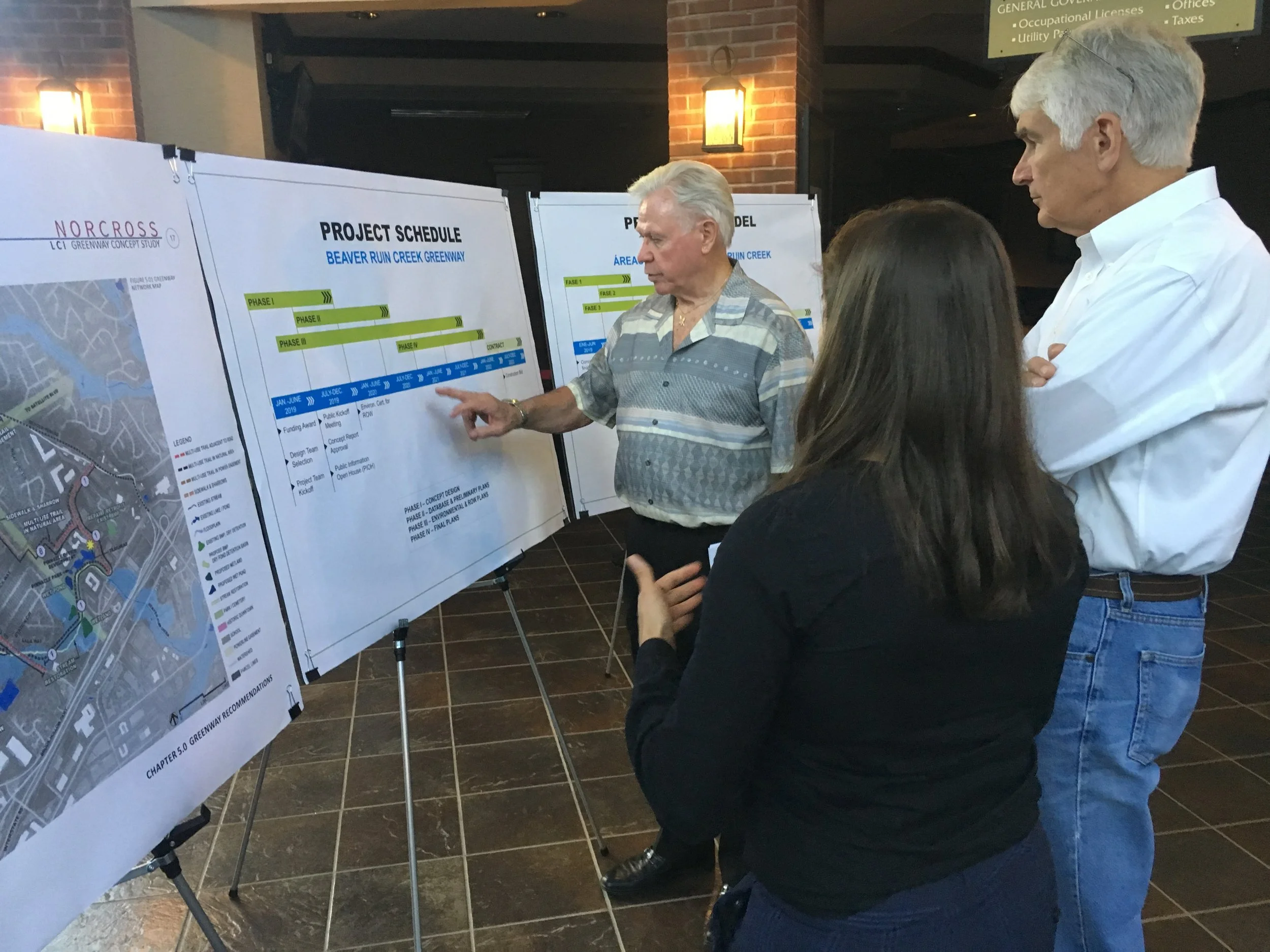 Three people examining project schedule posters at an indoor event, including a man pointing to details on a project schedule for Beaver Ruin Creek Greenway.