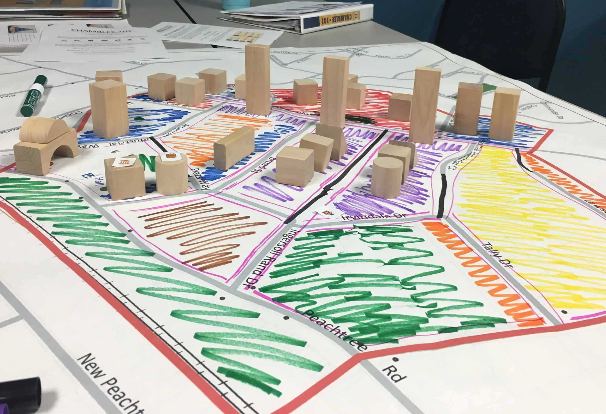 Colorful hand-drawn map of a neighborhood with streets and parks, featuring wooden blocks and shapes placed on various areas to represent buildings and landmarks.