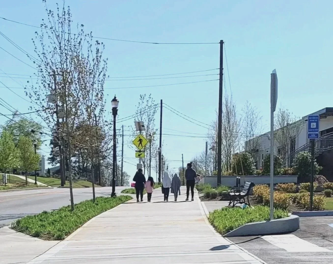 A sidewalk in a small town or city with four pedestrians, including two adults and two children, walking away from the camera. The scene includes trees, benches, streetlights, power lines, a pedestrian crossing sign, and a blue handicap parking sign, with clear weather and a bright blue sky.
