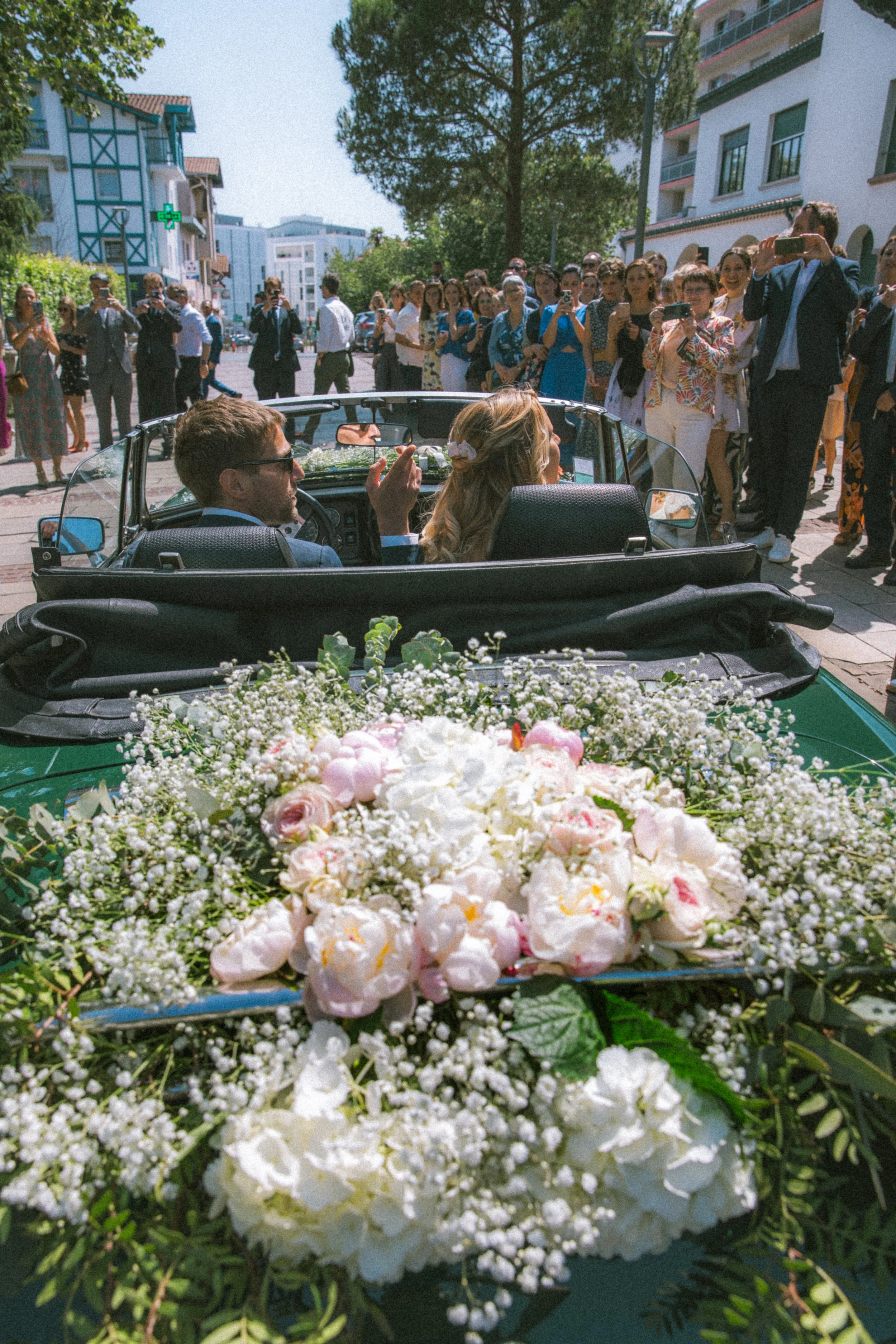 Un couple de mariés dans une voiture ancienne décorée avec des fleurs, entouré de nombreux invités qui prennent des photos à l'extérieur.