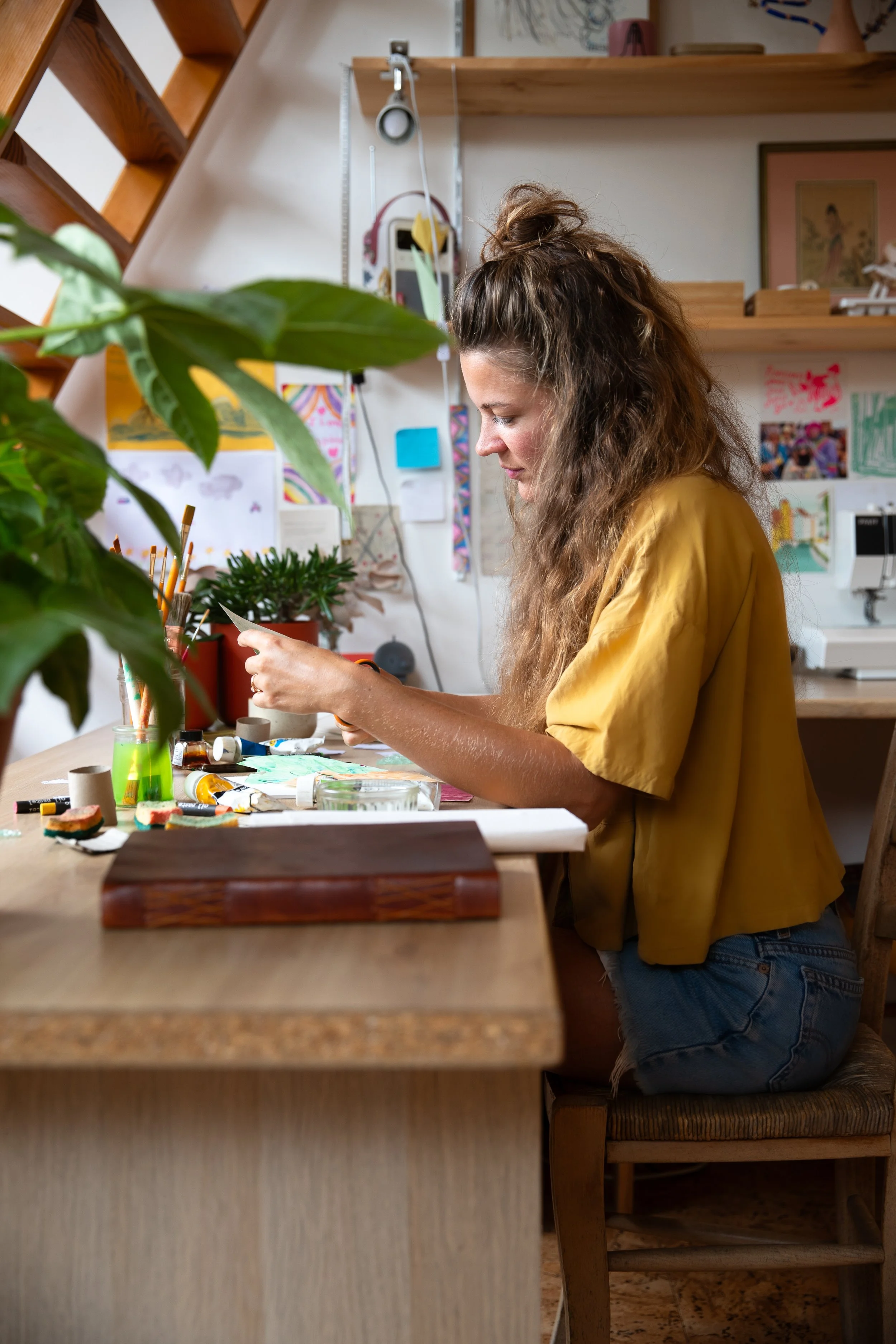 Jeune femme aux cheveux longs et bouclés, portant un t-shirt jaune, assise à une table en bois dans un atelier, travaillant sur un projet artistique avec de la peinture et des fournitures artistiques.