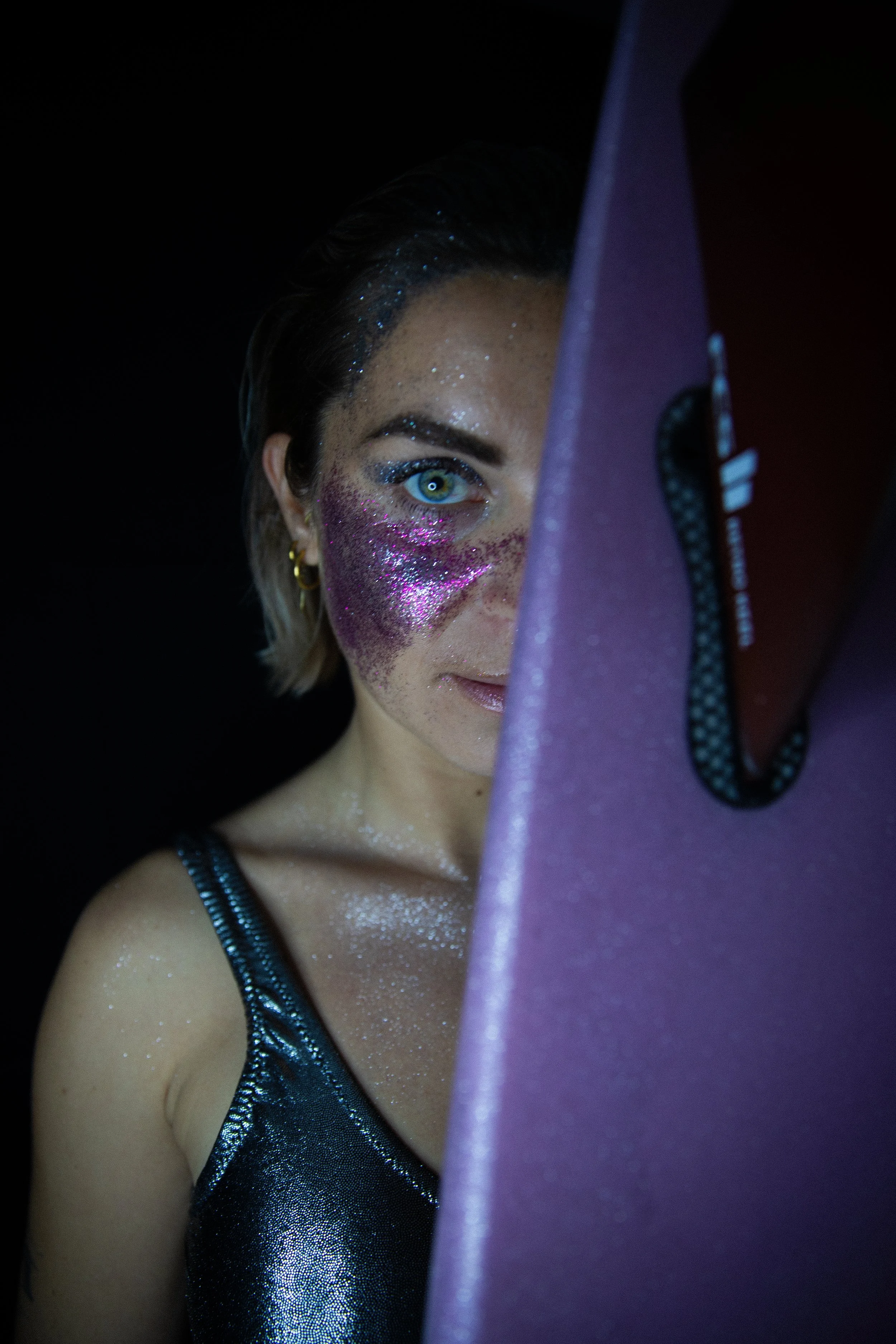 Portrait d'une femme avec un maquillage glittre violet et rose, regardant derrière un matériel de photographie, portant un vêtement noir brillant, fond noir.