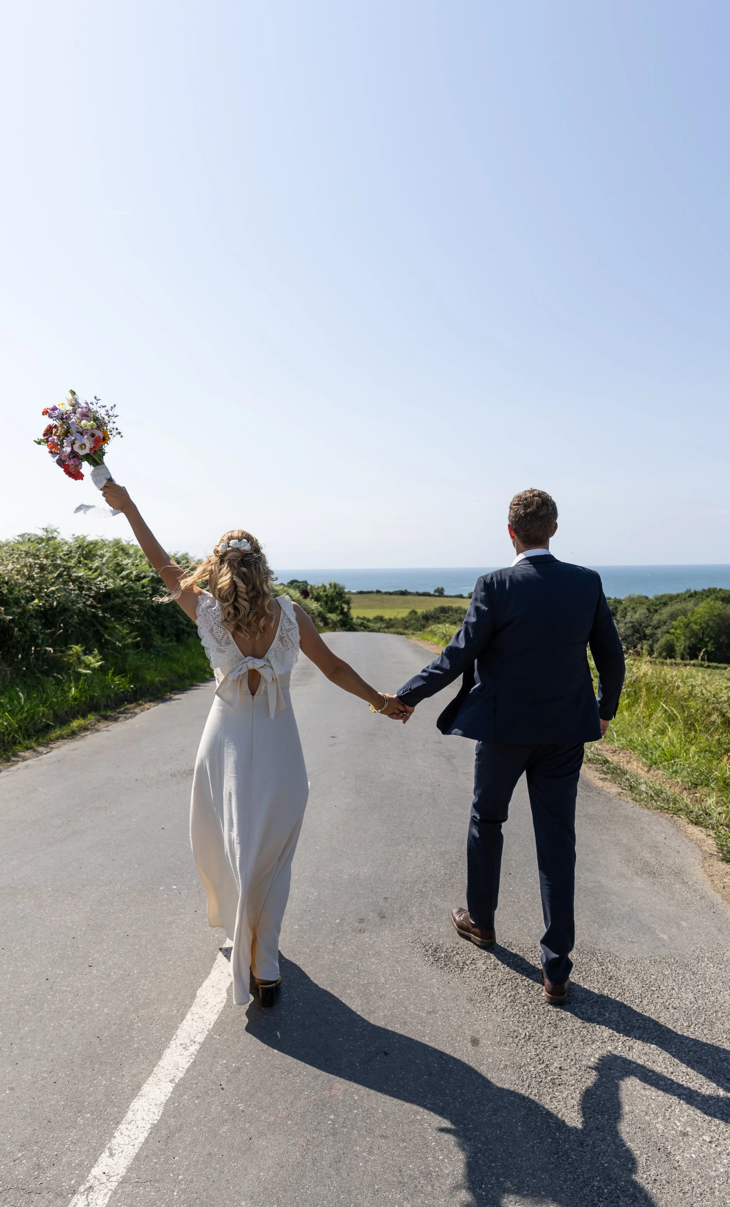 Un couple de mariés marche main dans la main sur une route vers la mer par une journée ensoleillée, la mariée brandissant un bouquet de fleurs
