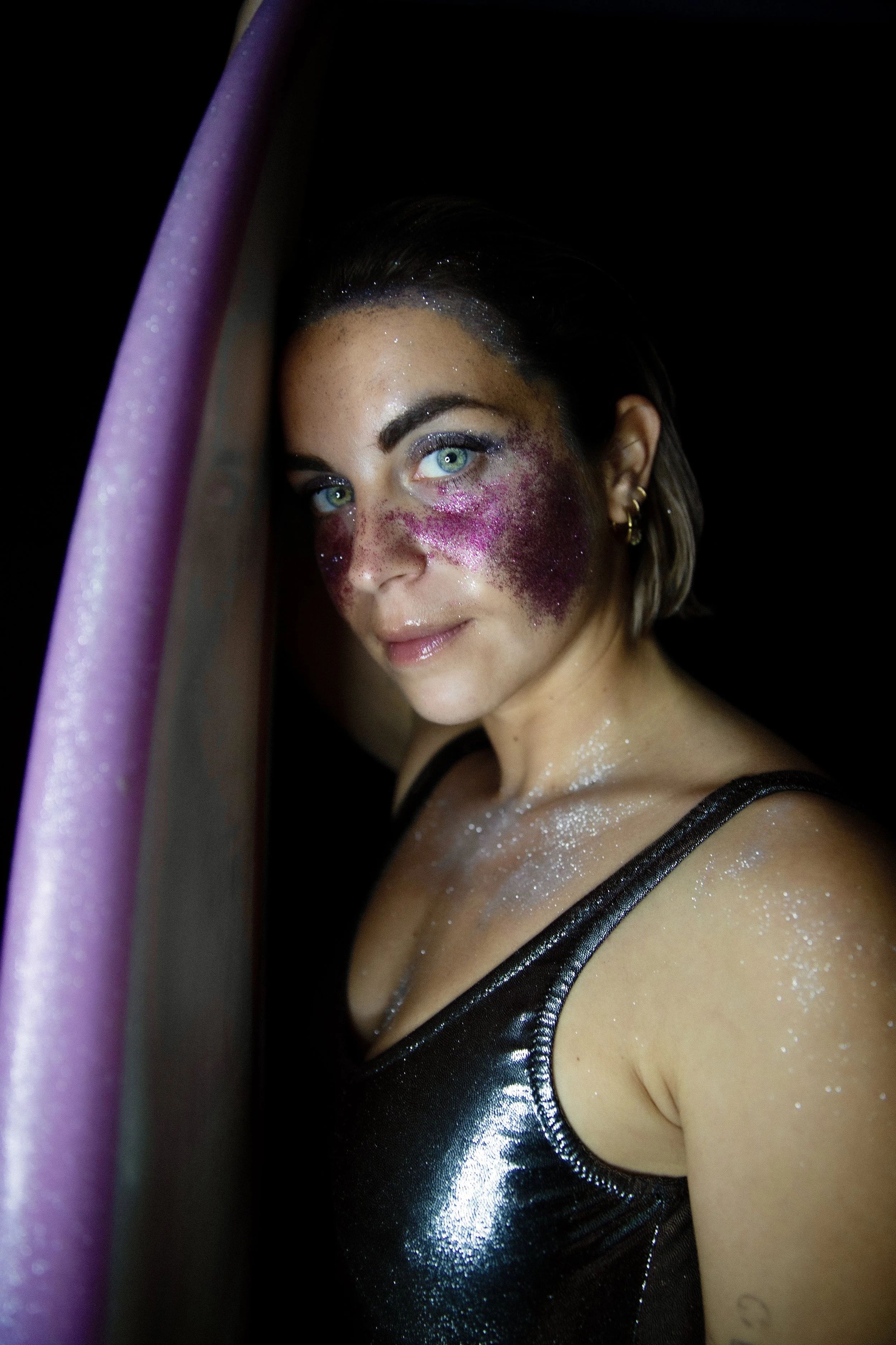 Une femme avec des yeux bleus, maquillage brillant et paillettes roses et violettes sur le visage, portant un haut noir en tissu brillant, regardant vers la caméra dans l'obscurité.