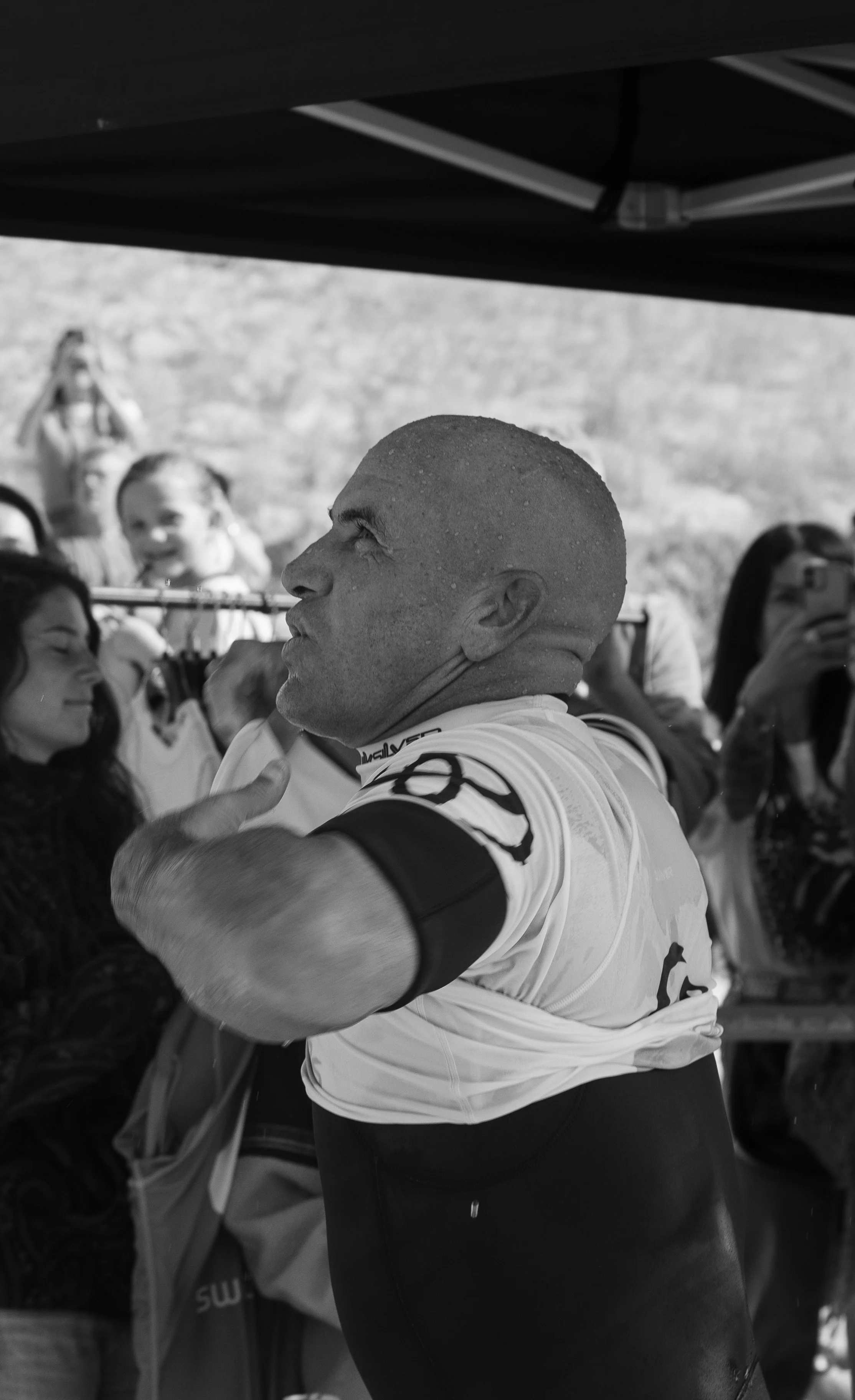 Un homme en maillot de cyclisme dans une scène de rassemblement ou compétition, entouré de plusieurs femmes et enfants qui prennent des photos ou regardent.