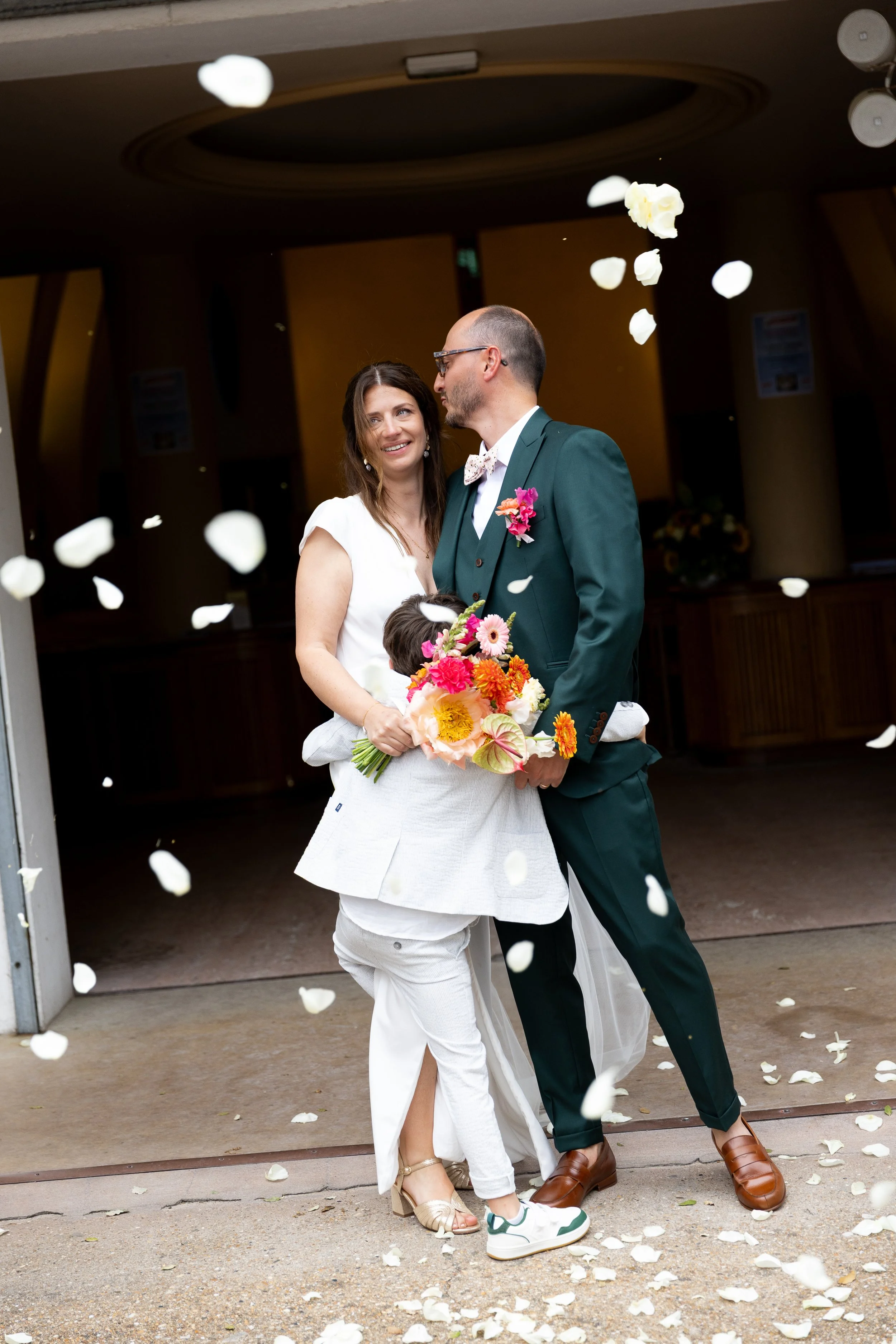 Un couple marié avec un enfant, tenant un bouquet de fleurs, sous des pétales de fleurs tombants, lors d'une cérémonie de mariage