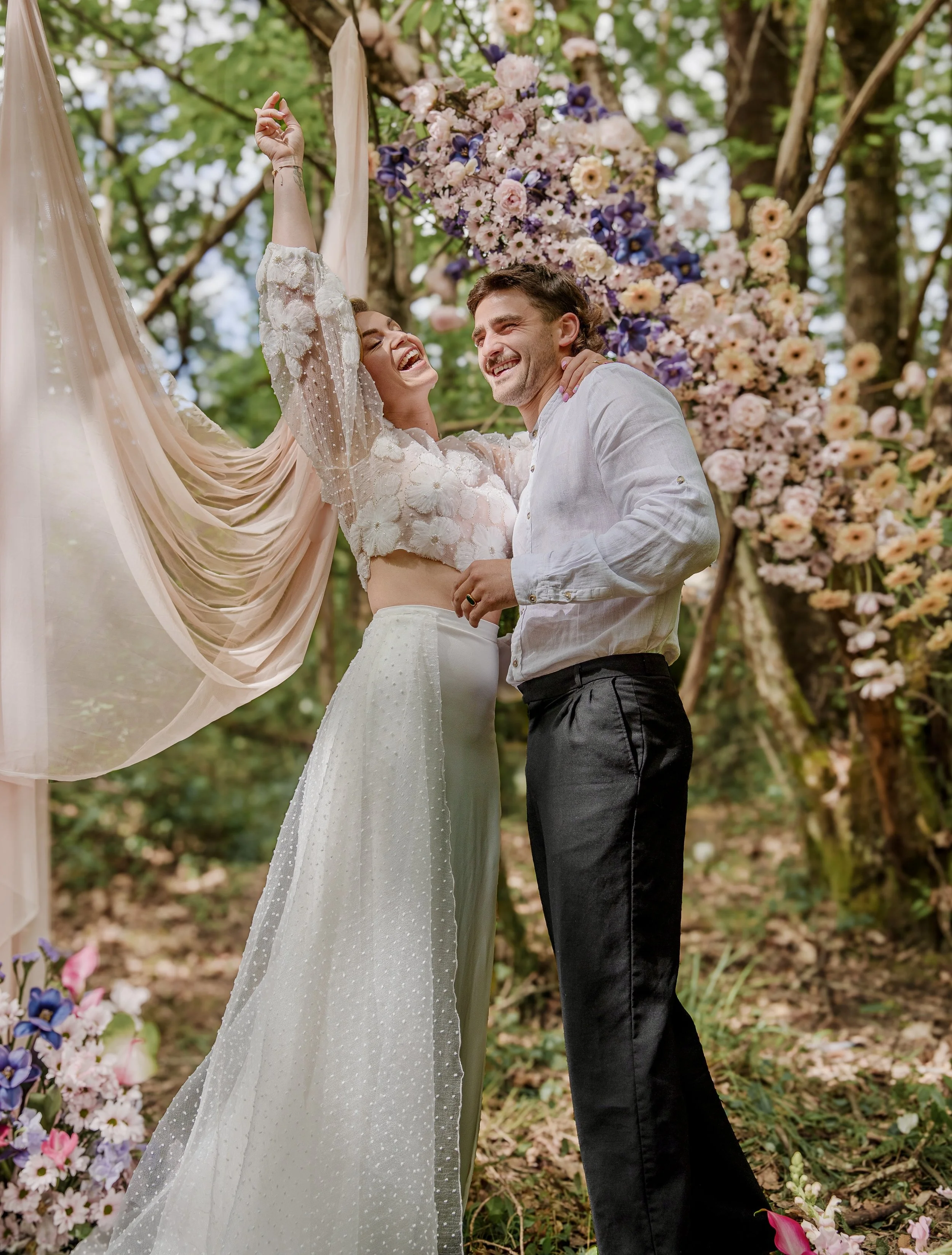 Un couple lors d'un mariage dans la forêt, souriant et dansant près d'une arche décorée de fleurs, avec des arbres en arrière-plan.
