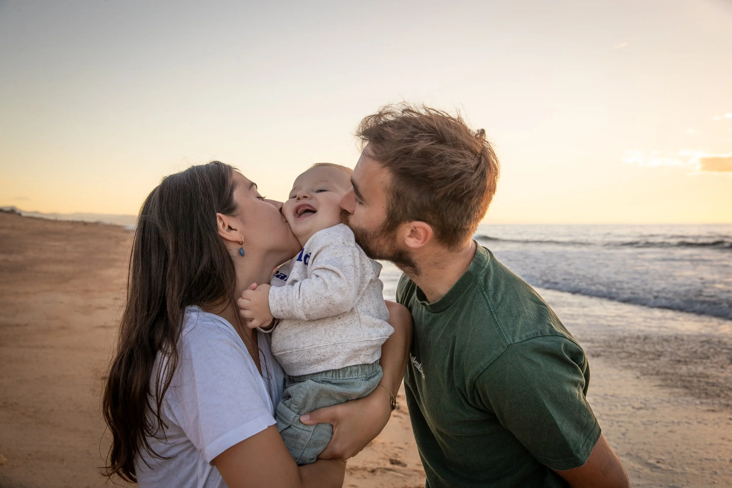 Une famille sur la plage au coucher du soleil, deux adultes embrassent leur bébé.