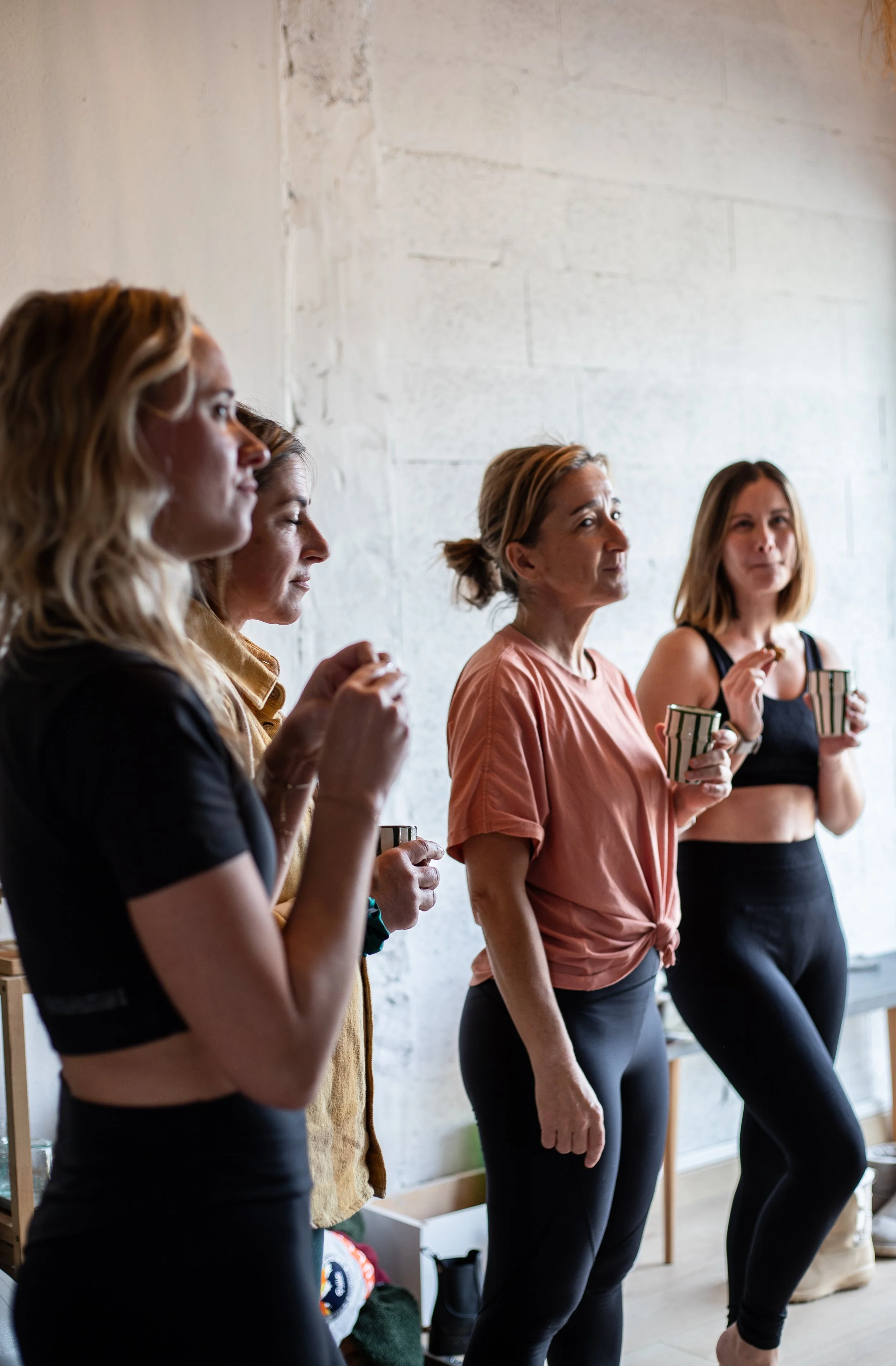 Cinq femmes en tenue de sport debout contre un mur en brique blanche, tenant des tasses, semblant écouter ou participer à une conversation ou une activité de groupe.