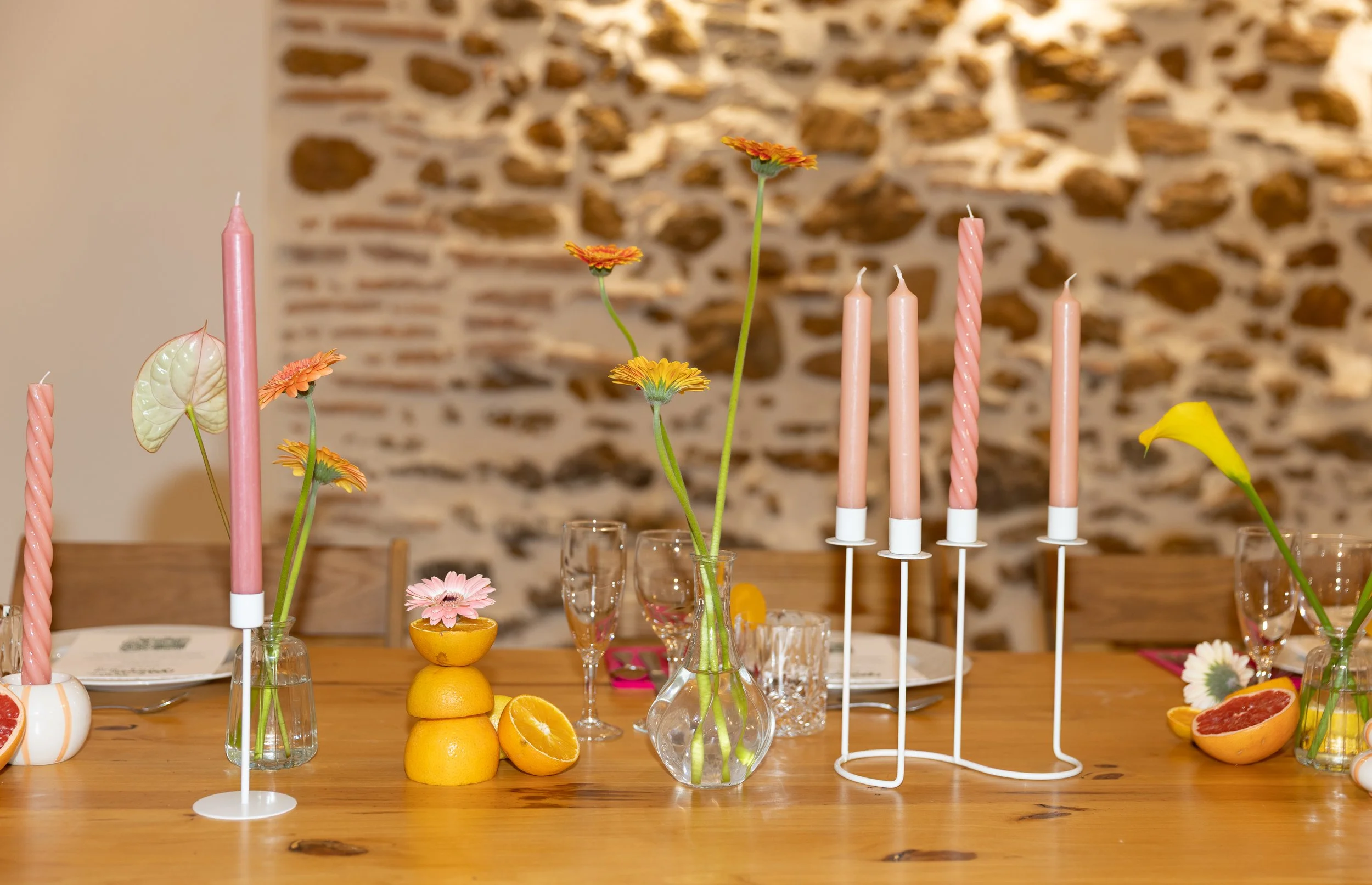 Décoration de table avec bougies roses, fleurs colorées dans des vases, citrons, pamplemousses, anthurium jaune, et éléments floraux, en arrière-plan mur en pierre.