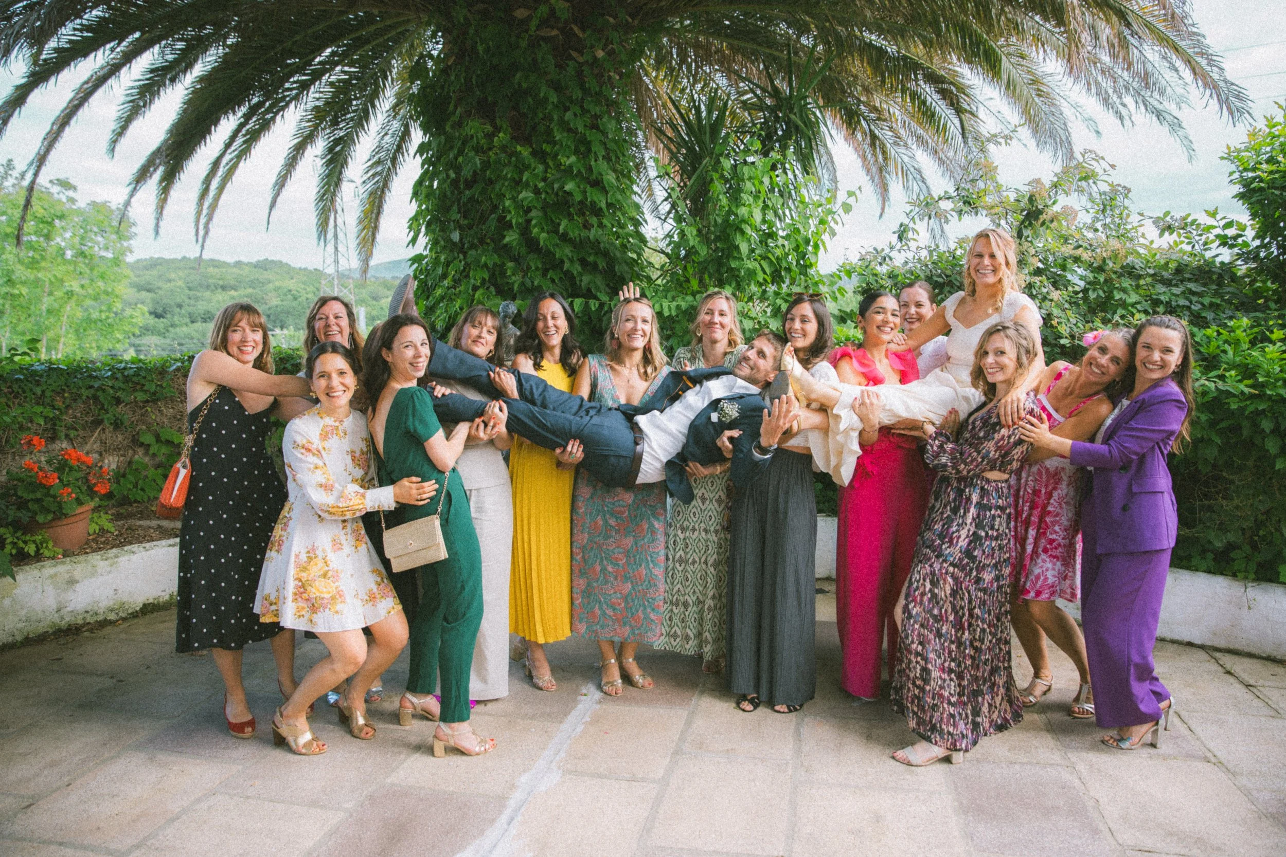 Groupe de personnes souriantes prenant la pose lors d'une célébration en extérieur, avec une femme en robe blanche portée par plusieurs amis.