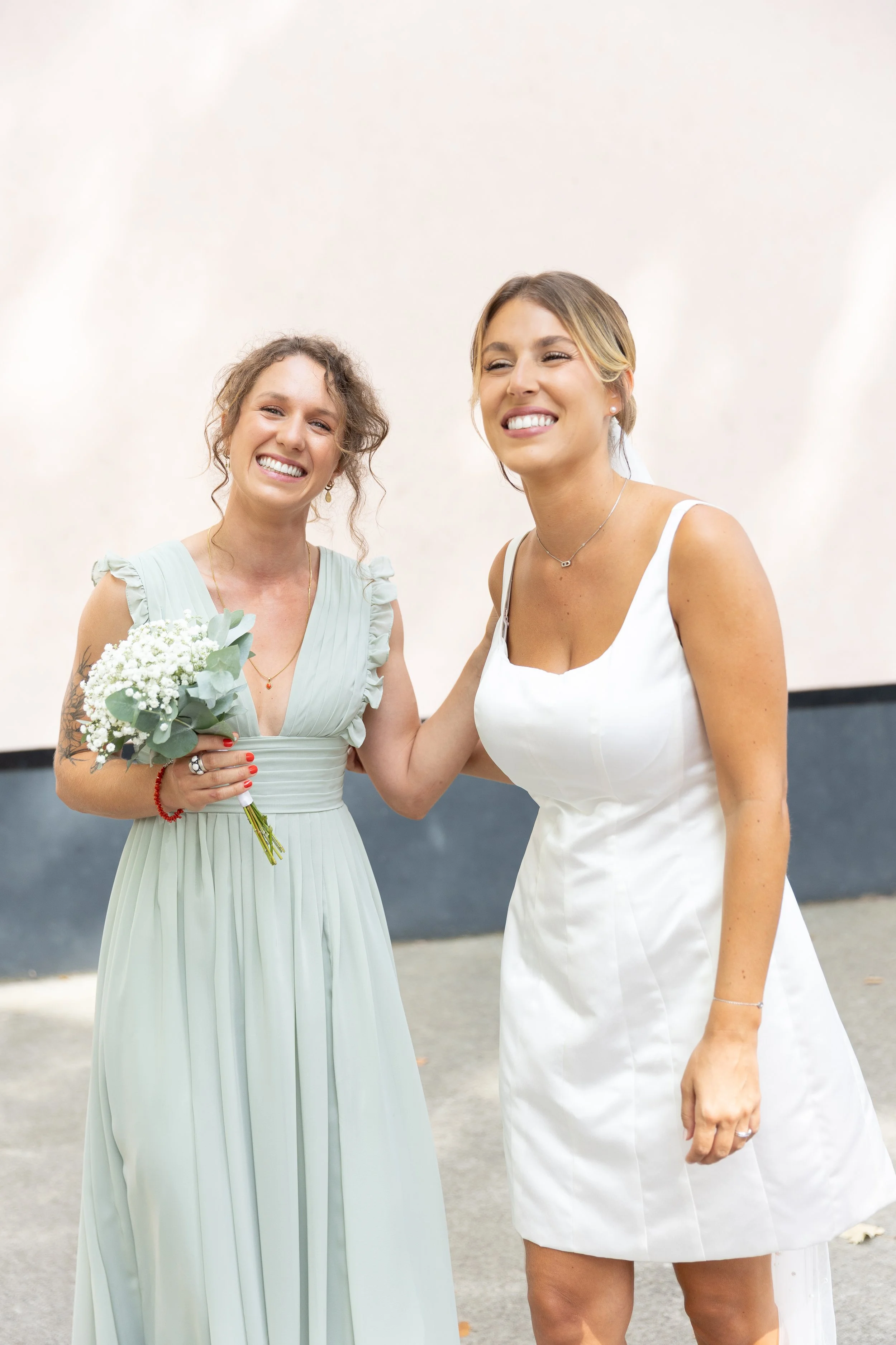Deux femmes souriantes, l'une en robe blanche de mariée, l'autre en robe couleur pastel, se tenant par l'épaule lors d'un mariage en extérieur.