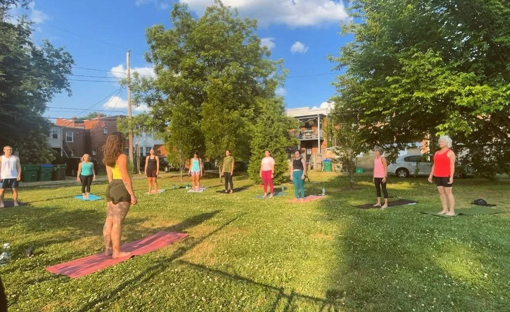 Group of people practicing yoga outdoors on a grassy area with trees and houses in the background, under a blue sky.