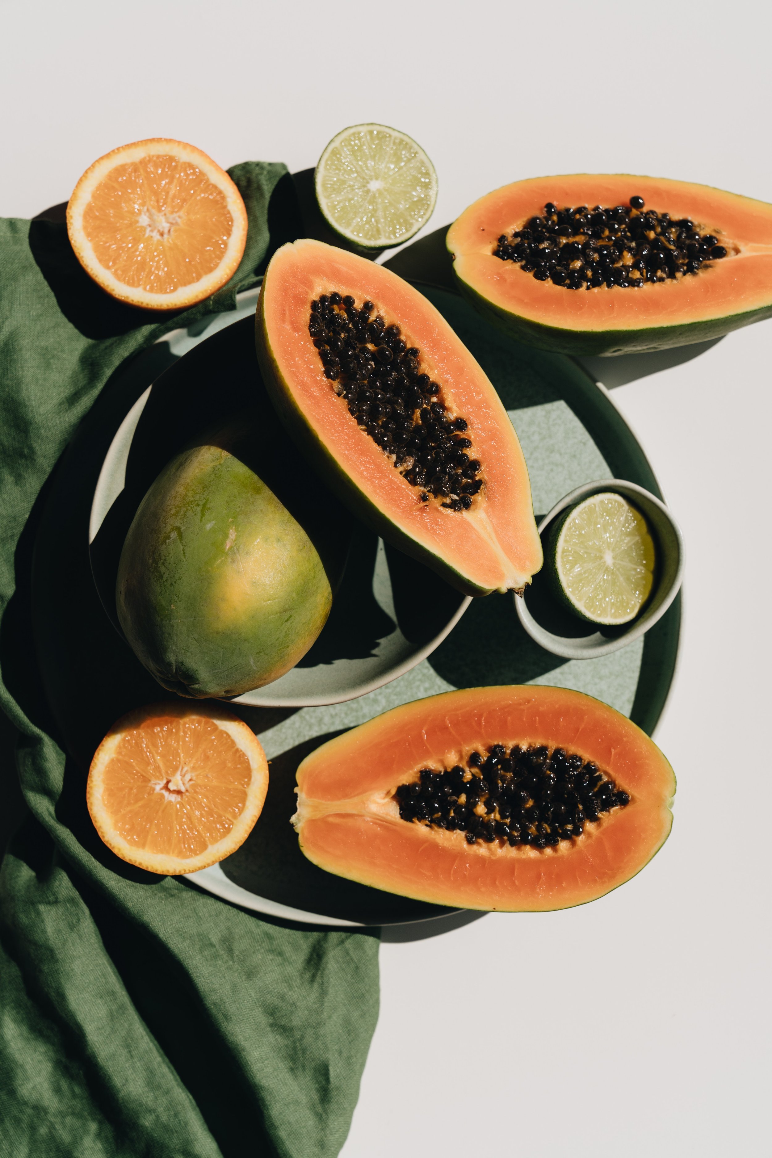 Halved papayas with black seeds, halved oranges, and lime slices arranged on a green cloth and a white plate.
