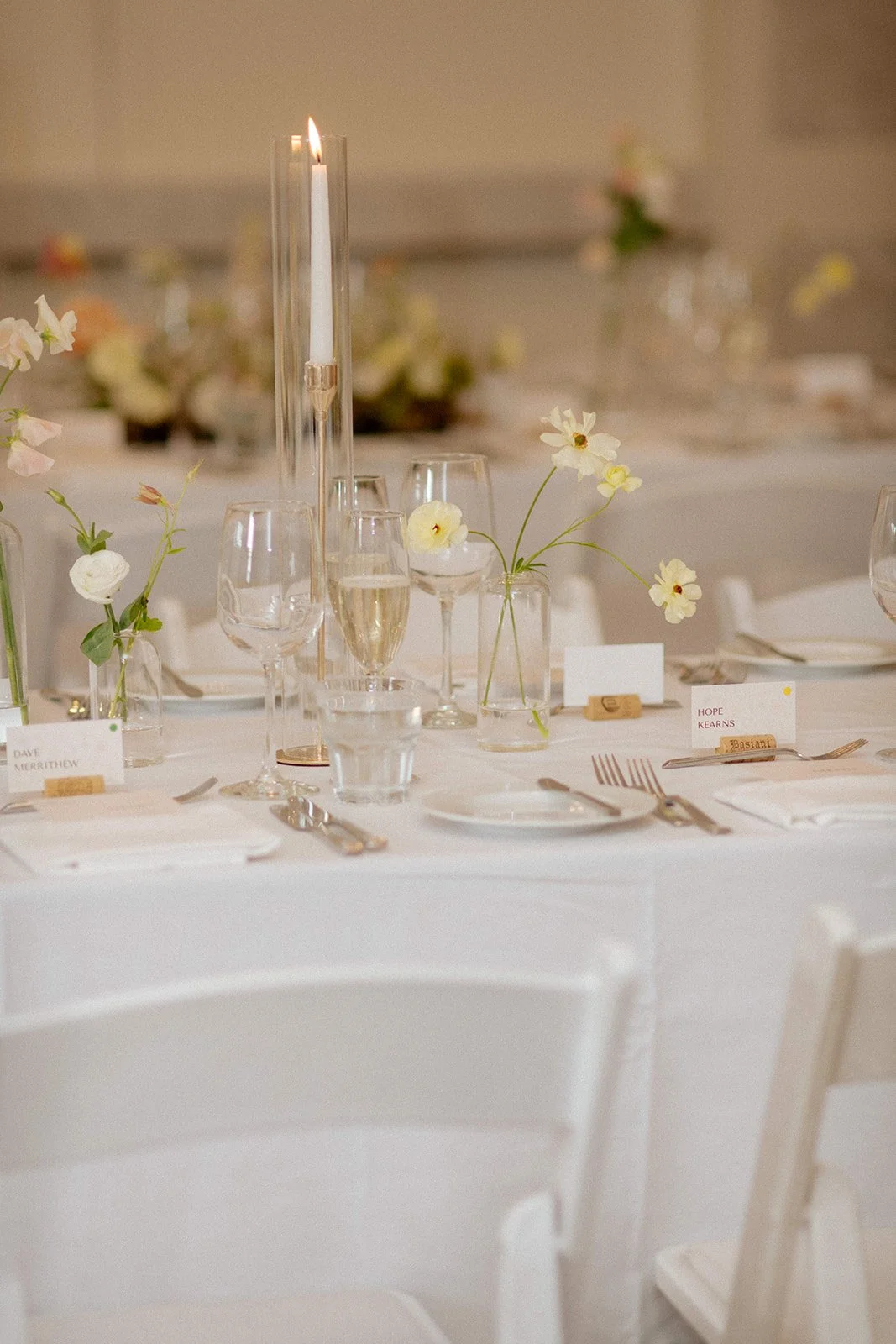 Elegant table setup for a wedding or formal event with white tablecloths, white chairs, floral centerpieces with white flowers in glass vases, champagne and water glasses, and place cards. Top wedding venues toronto