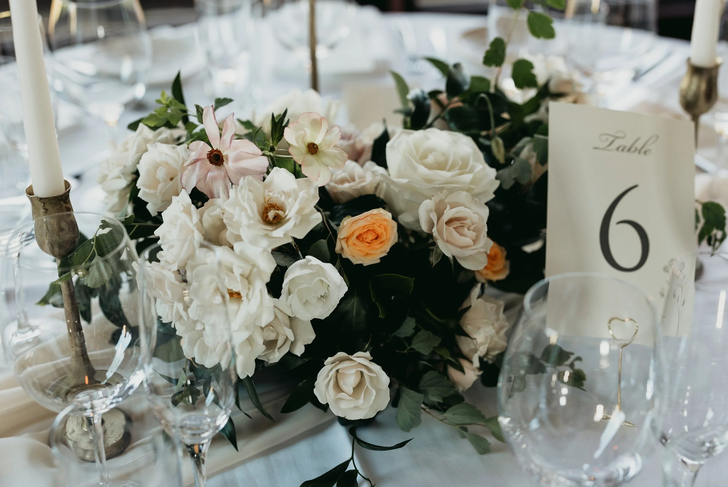 Elegant wedding table centerpiece with white, blush, and peach flowers and greenery, with a table number 6 card, clear glassware, and white napkin.