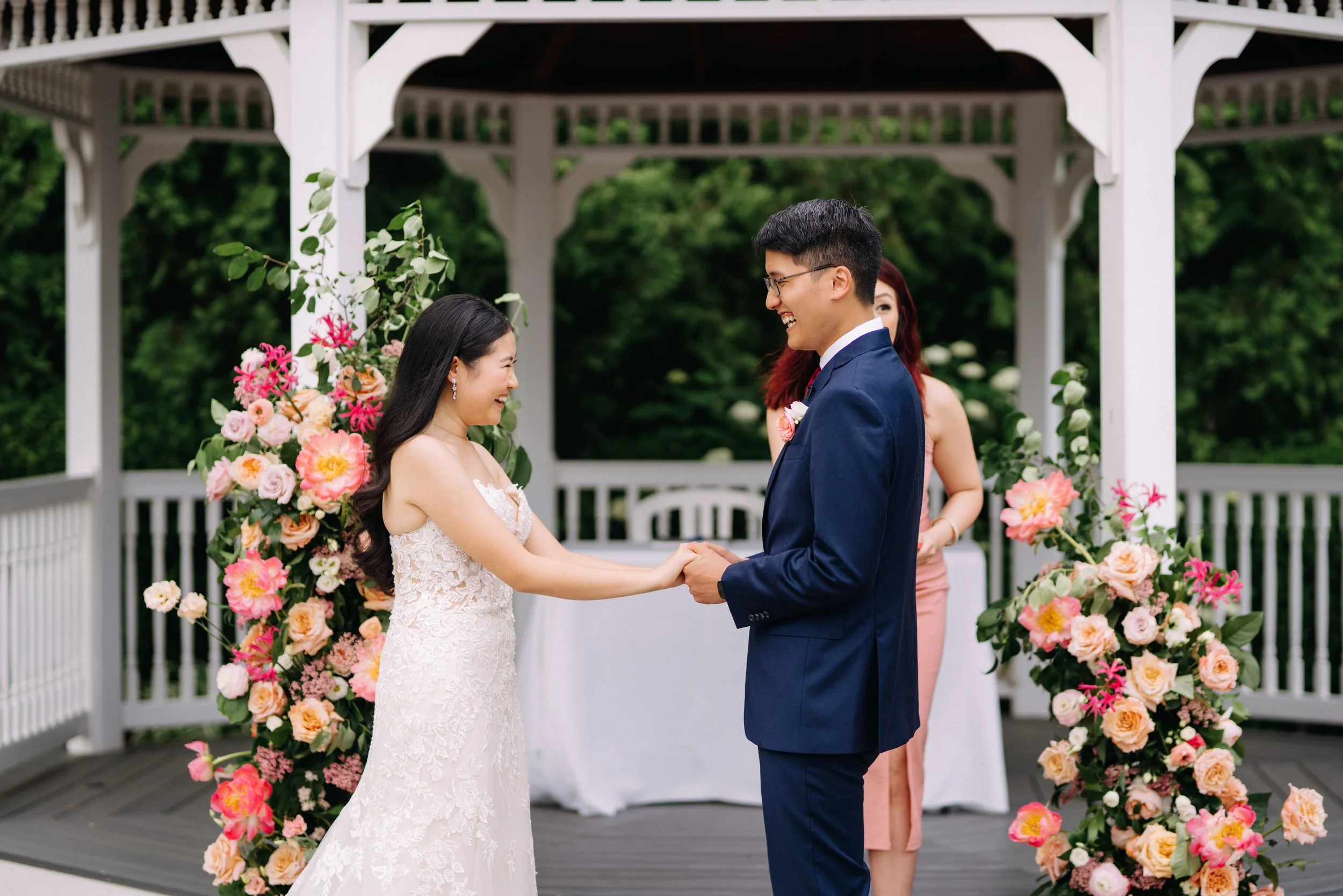 A bride and groom holding hands and smiling at each other during their outdoor wedding ceremony, with floral arrangements and a white gazebo.