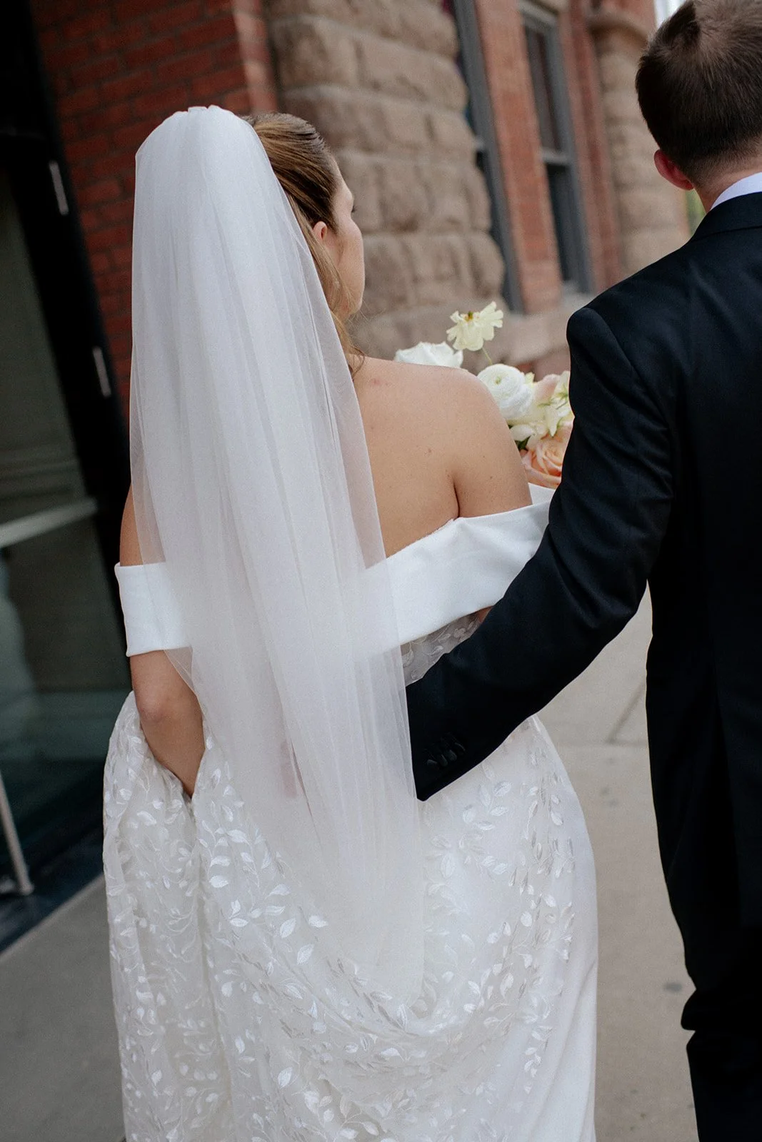 A bride and groom standing outside, holding a bouquet of flowers, with the bride wearing a white off-shoulder wedding gown and a long veil, and the groom in a black suit. The scene appears to be at a wedding ceremony. Top wedding florist