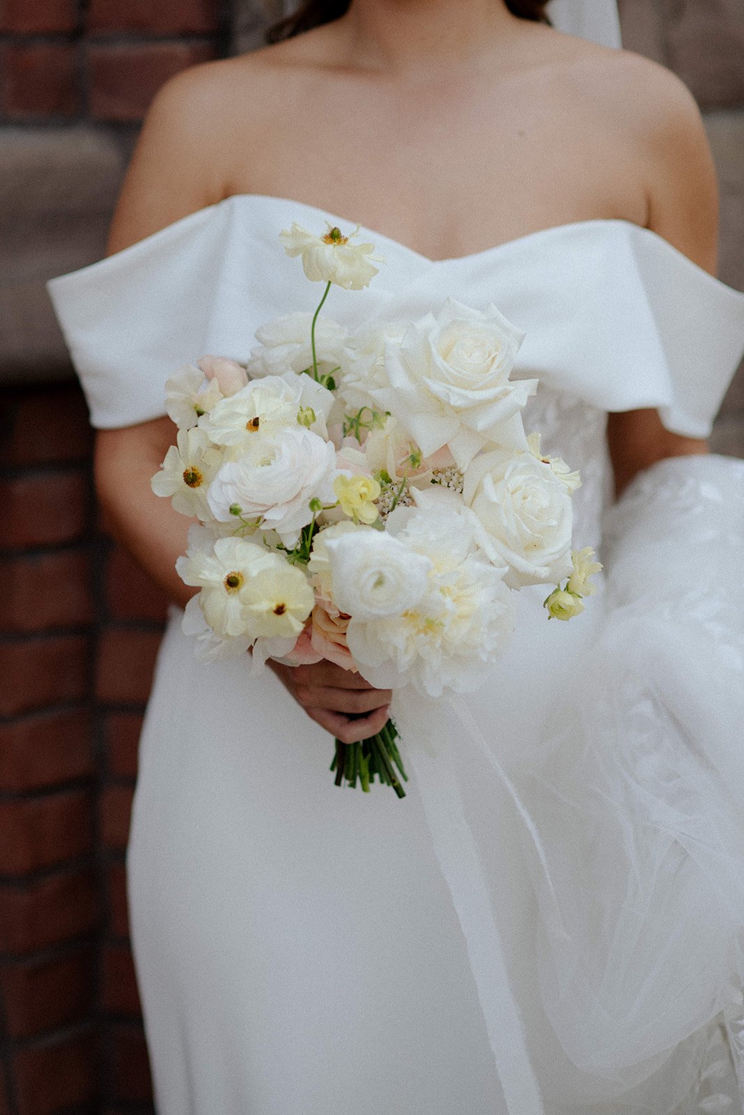 A bride in a white wedding dress holding a bouquet of white and pale pink flowers, standing near a brick wall. Best local flower wedding toronto