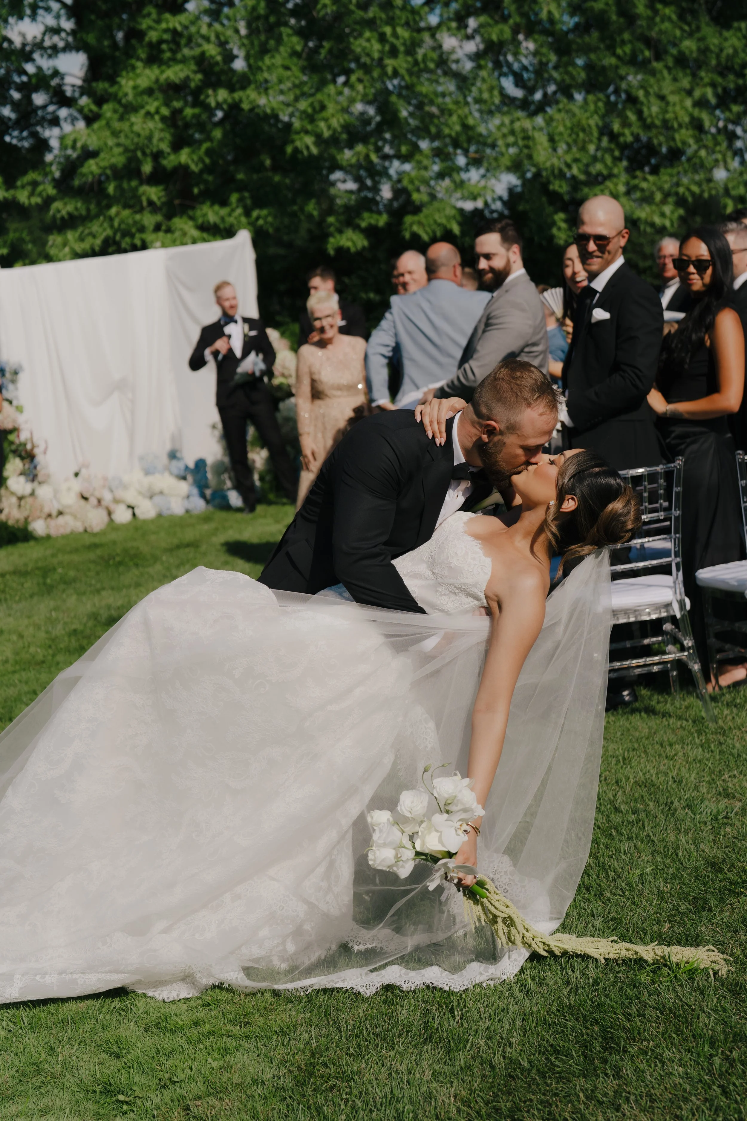 A bride and groom are kissing during their outdoor wedding ceremony, with wedding guests seated around them and a floral backdrop in the background.