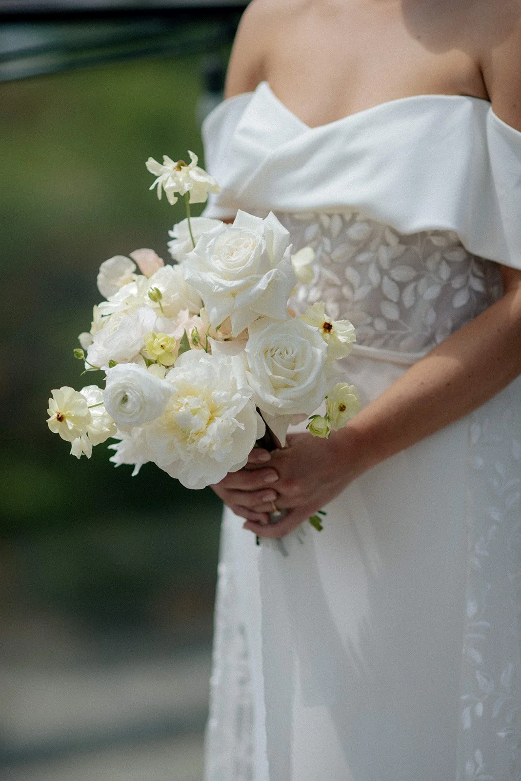 A bride holding a bouquet of white roses and light cream flowers, wearing a white wedding dress with floral embroidery and an off-the-shoulder sleeve. Top wedding florist Toronto