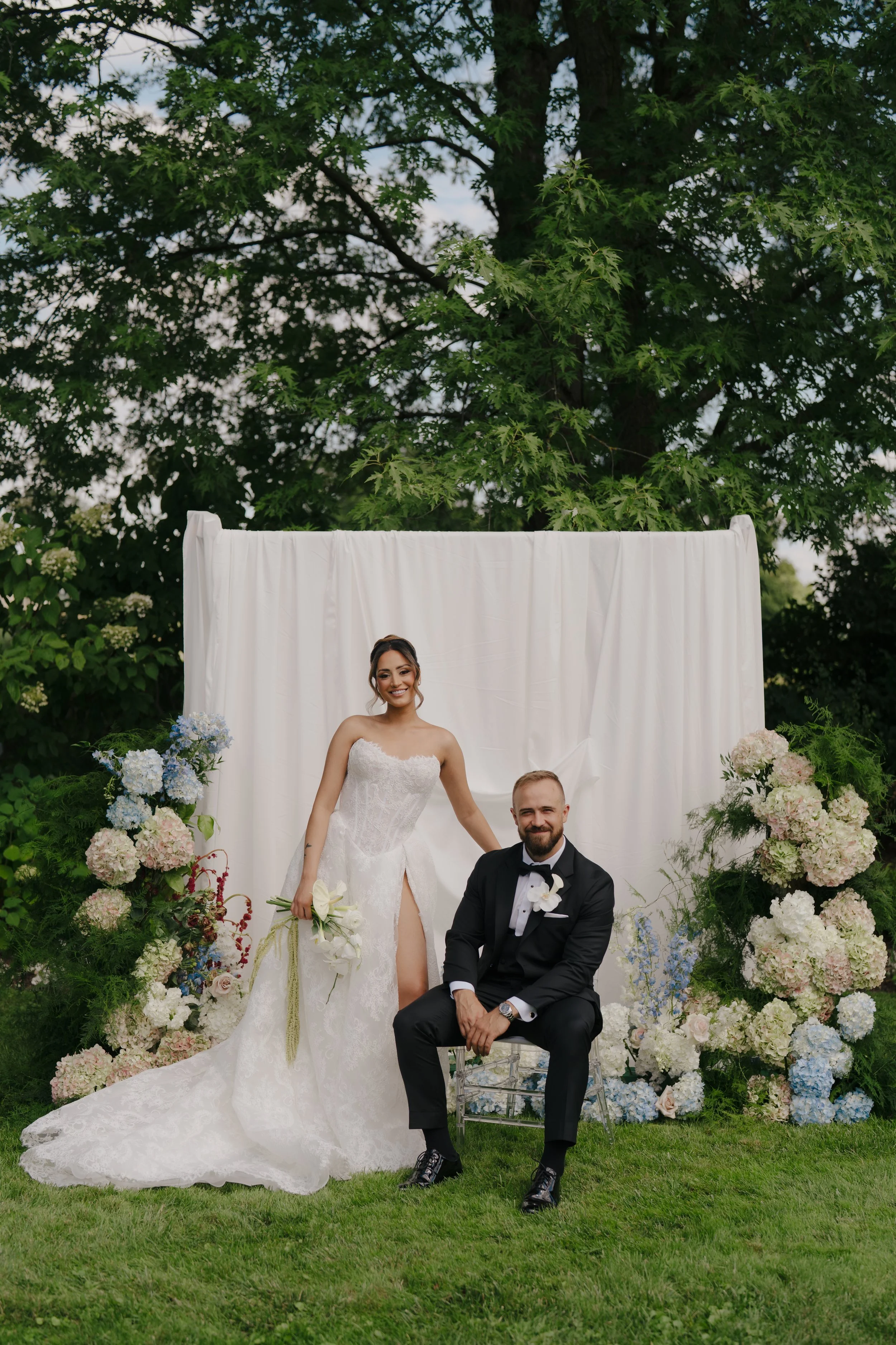 Bride and groom posing outdoors for their wedding photo, with the bride standing and the groom sitting on a transparent chair, surrounded by white and pastel flowers, and a white backdrop, in a lush green garden.