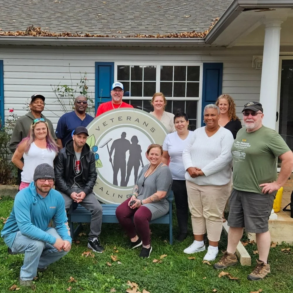 Veterans and volunteers gathered around the Veterans Healing Farm sign in front of the farmhouse