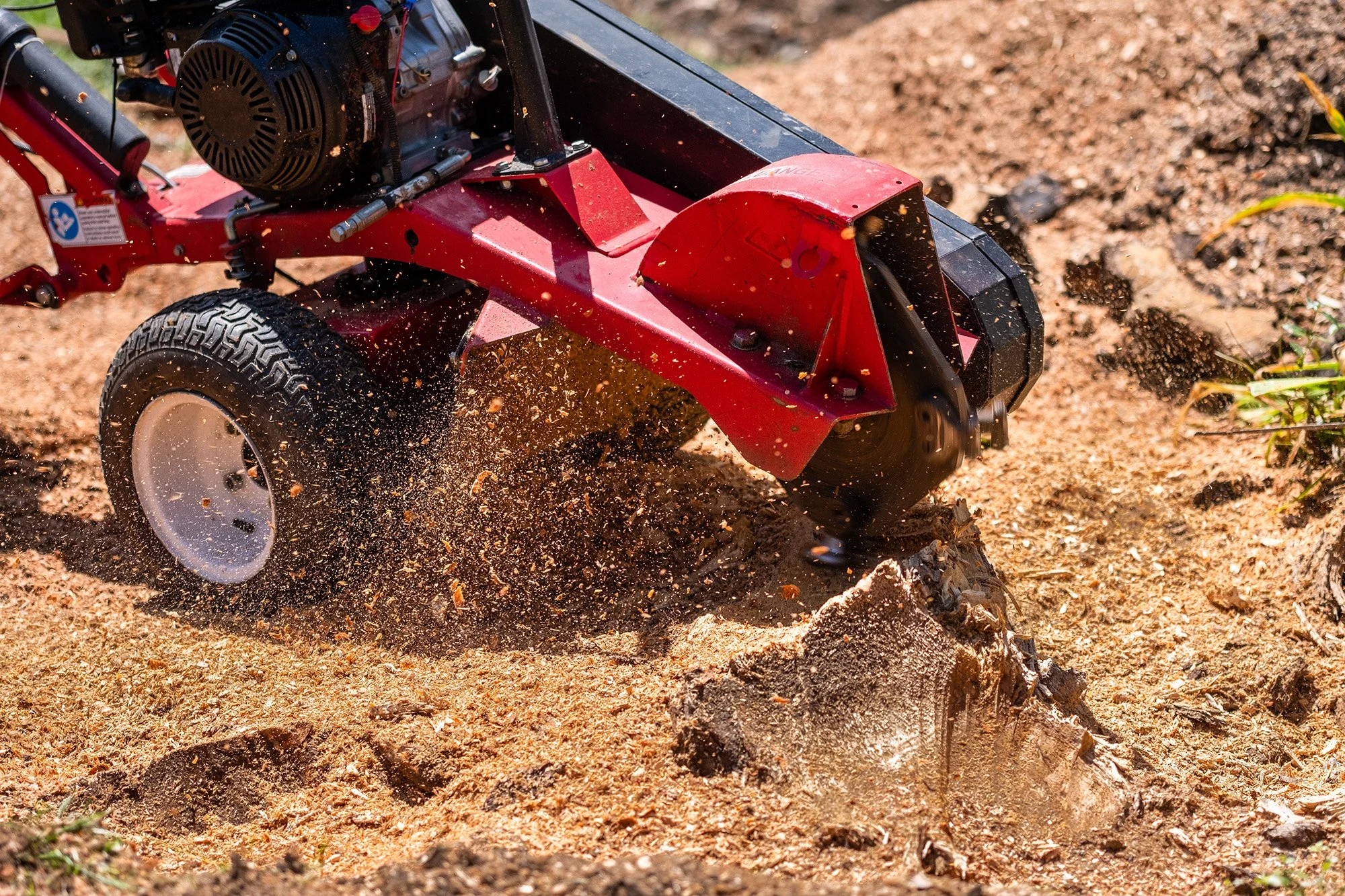 A red and black rotary tiller attachment working on soil, with dirt and small rocks flying as it tills the ground.