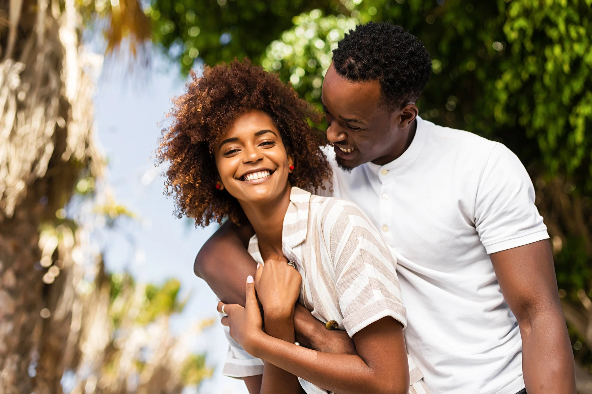Happy young couple laughing outdoors, man embracing woman from behind under palm trees on a sunny day.