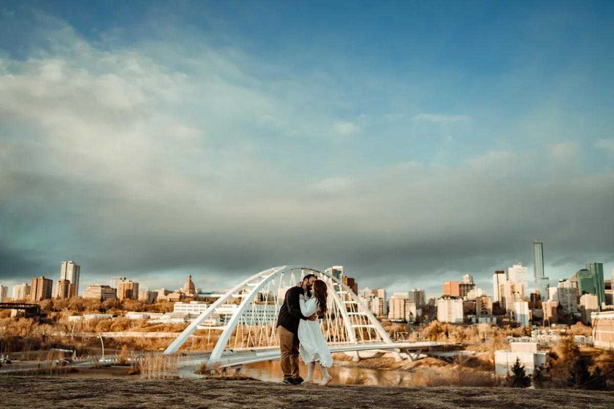 A couple share a romantic kiss while being framed in Edmonton's Walterdale bridge with the city in the background.