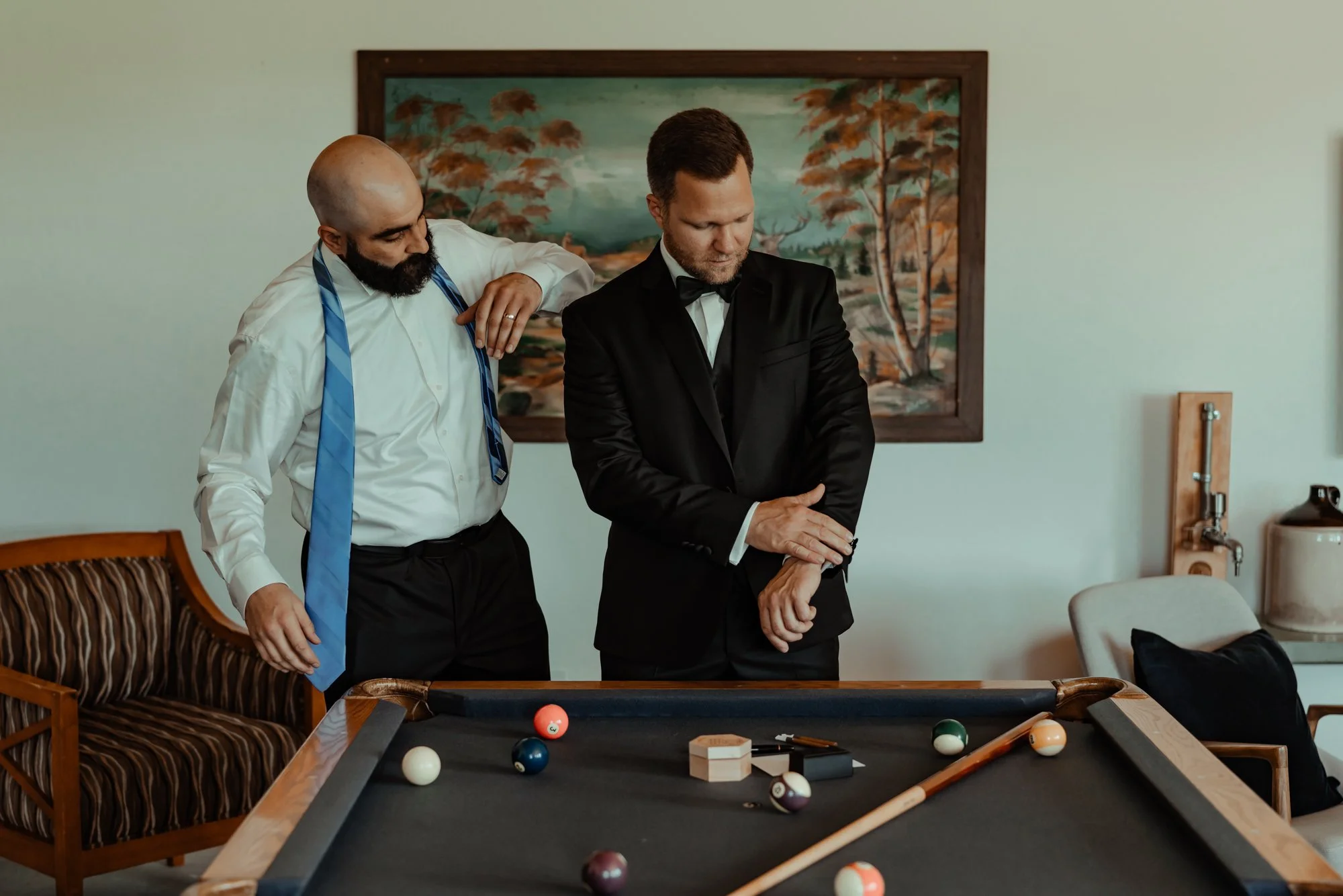 A best man helps the groom into his tuxedo in front of a pool table.