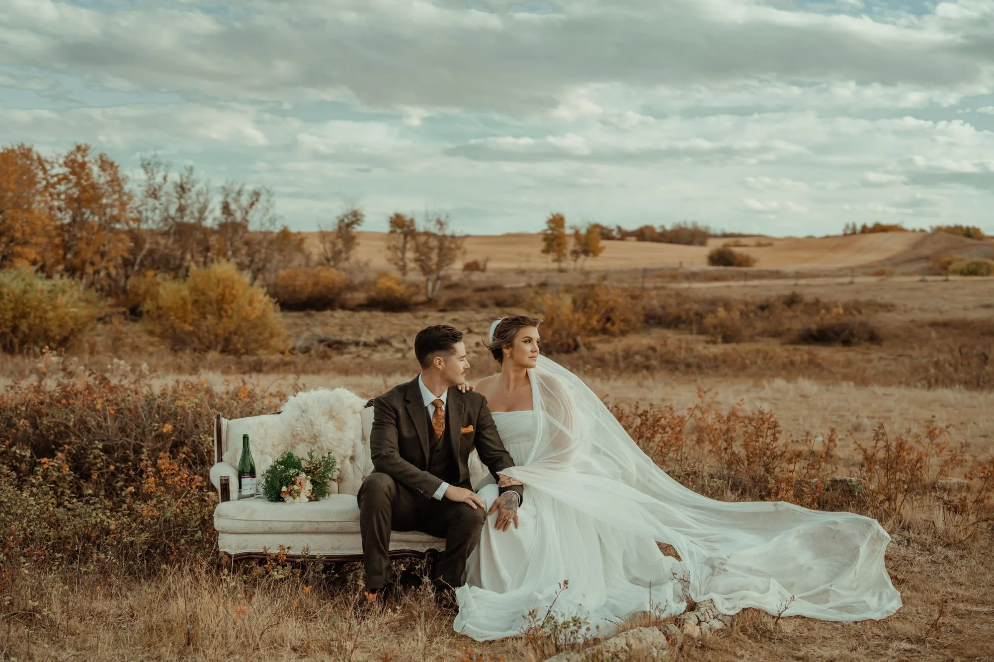 A bride and groom pose for an editorial portrait while sitting on a vintage couch with sparkling wine in a prairie field