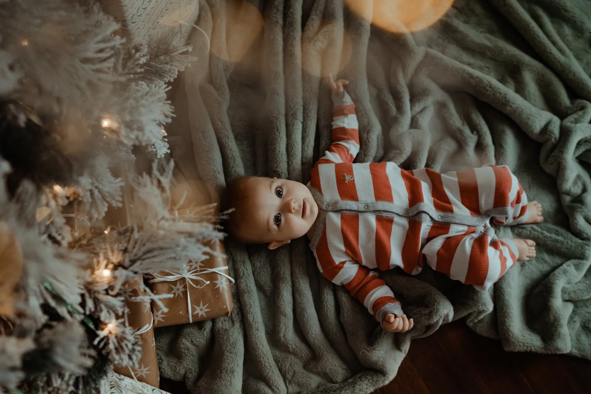 Baby lying on a soft, green blanket next to a decorated Christmas tree with wrapped gifts underneath, illuminated by warm lights.