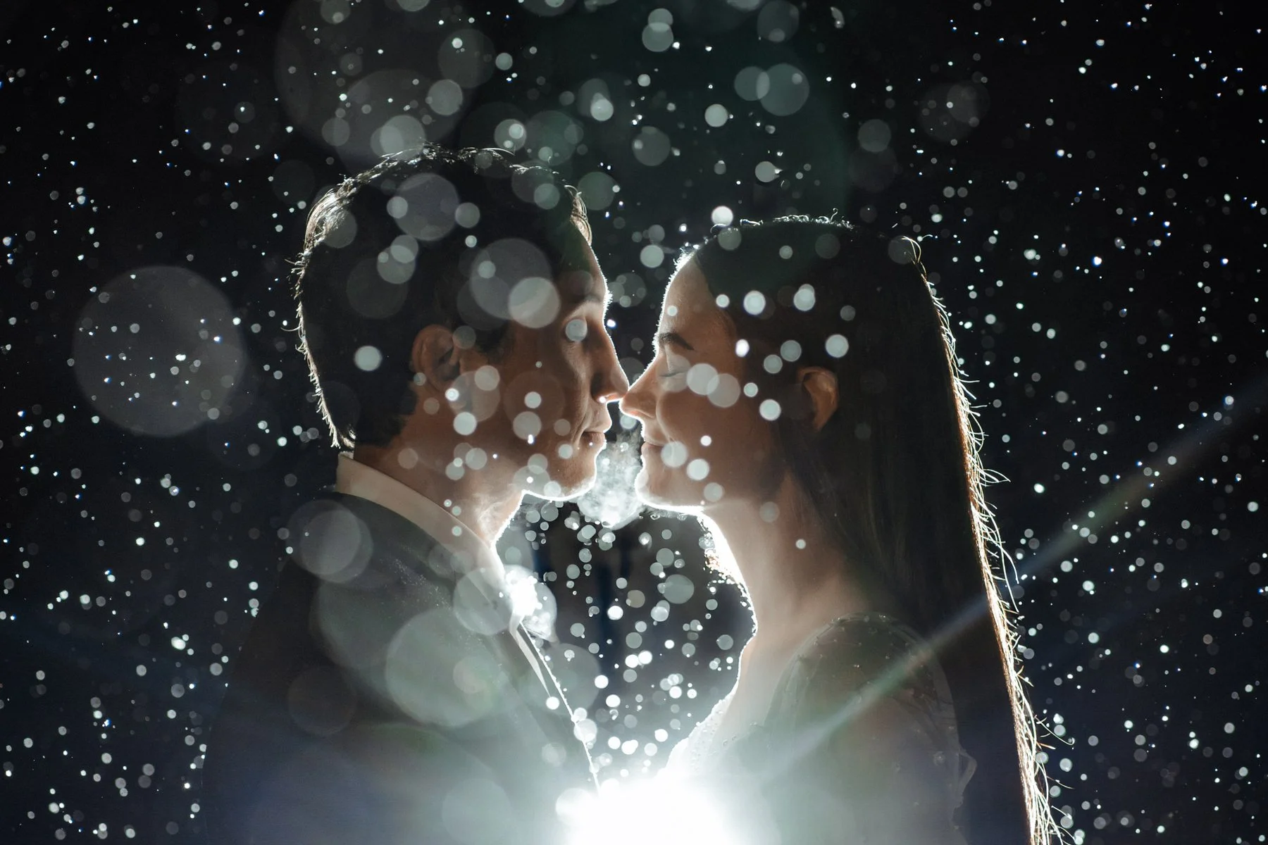 Snowflakes fall around a bride and groom who are leaning in to kiss.