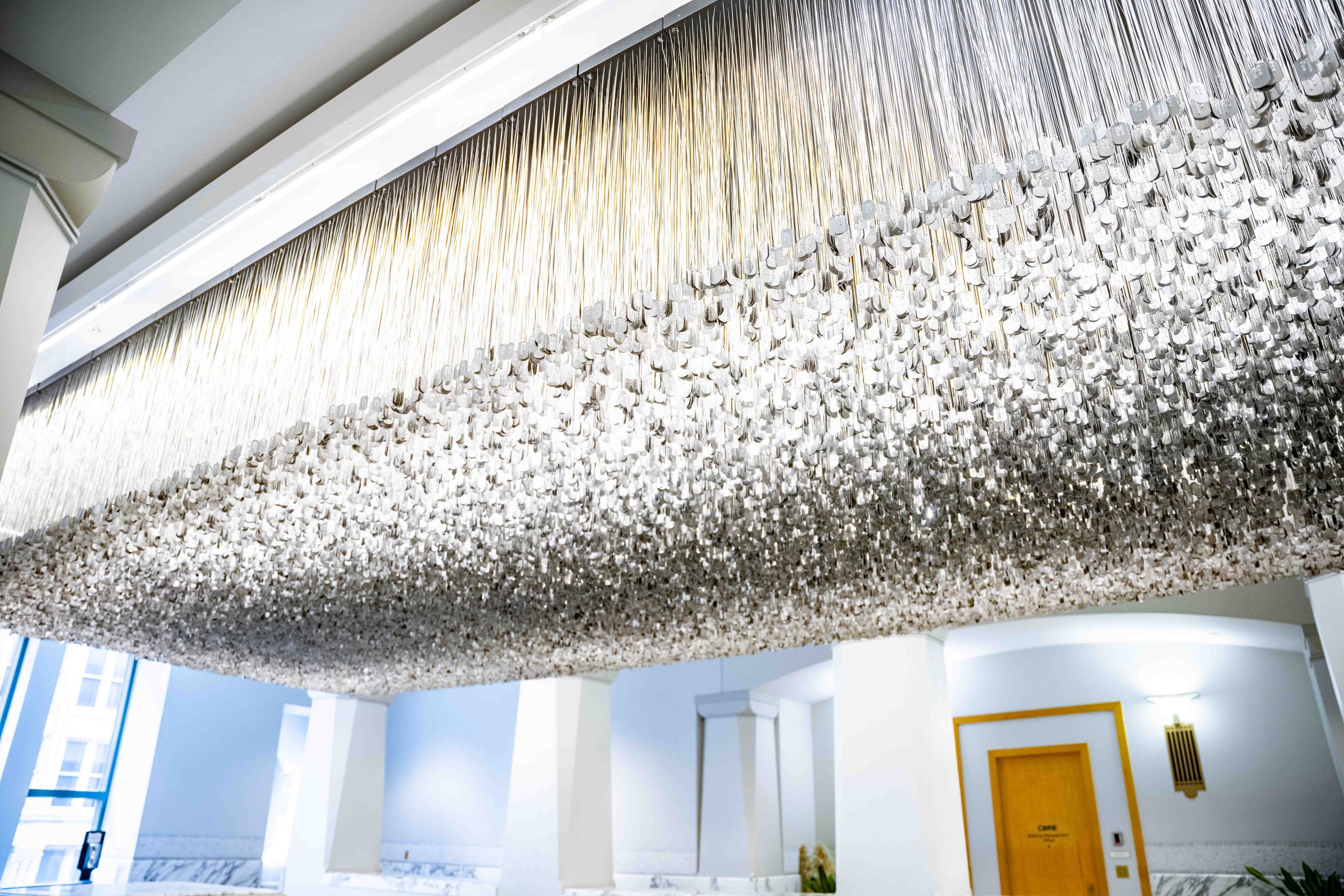 58,307 stainless steel dog tags suspend from a 10 foot by 40 foot space in the ceiling of the third floor of the Harold Washington Library Center. Underneath are escalators. The space has white walls, and tall windows.