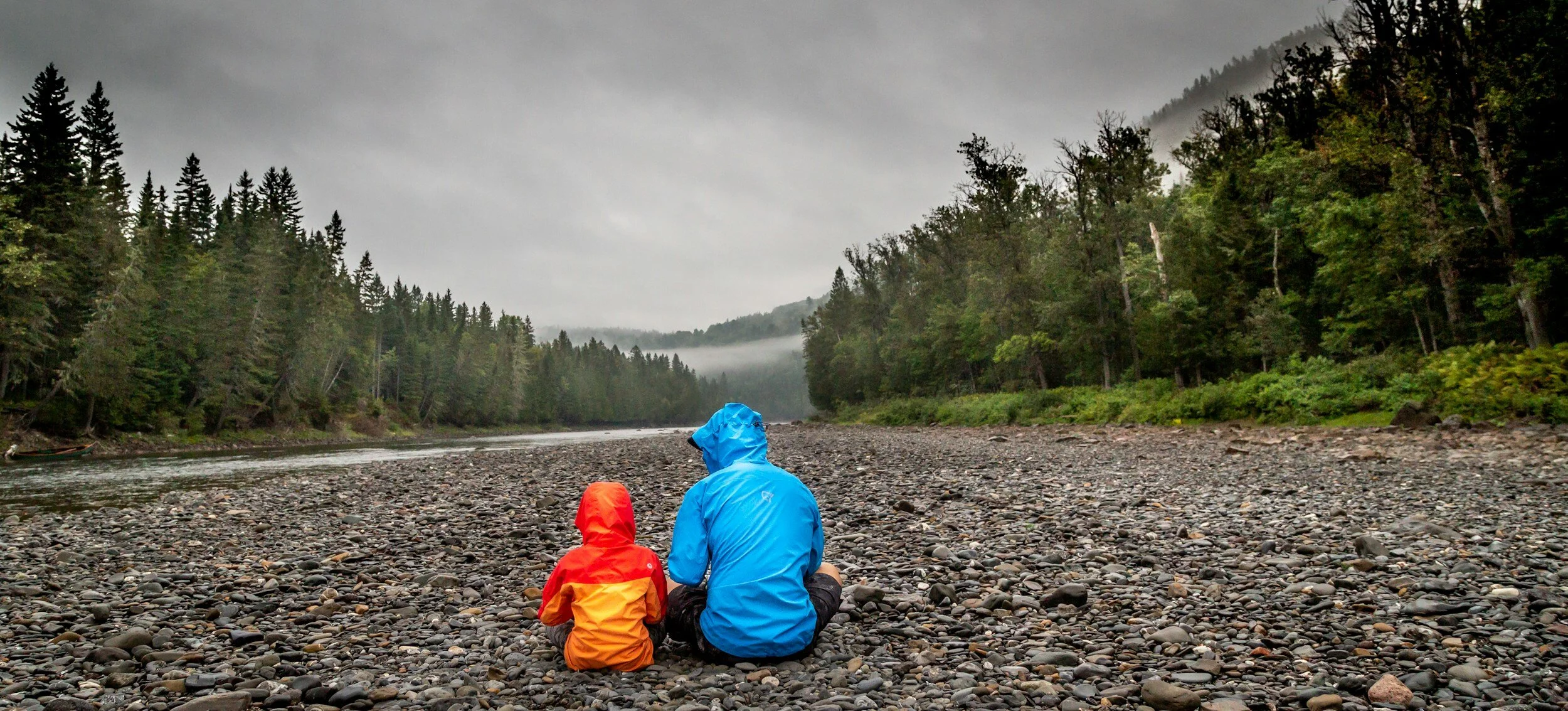 Two people wearing rain jackets sitting on a rocky riverbank, surrounded by forest trees under a cloudy sky.