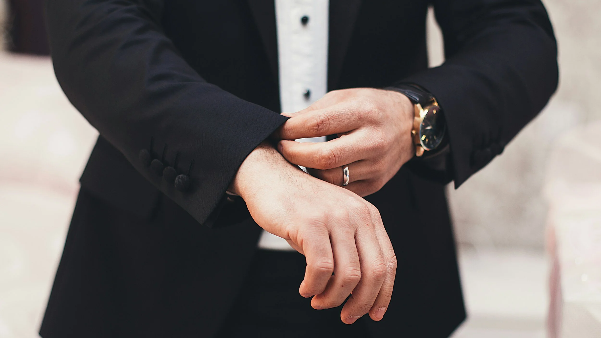 Man in a black suit adjusting sleeve, wearing a watch and a ring.