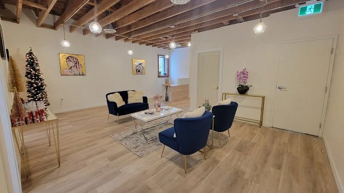 Modern living room with wooden floors, blue chairs, a white couch with cushions, a coffee table, and decorative artwork. Christmas tree with gold ornaments on the side, and hanging lights overhead.