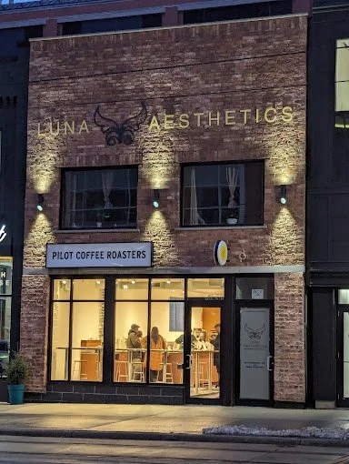 Exterior view of Pilot Coffee Roasters with Luna Aesthetics signage, showing a brick facade, large windows, and interior seating area.