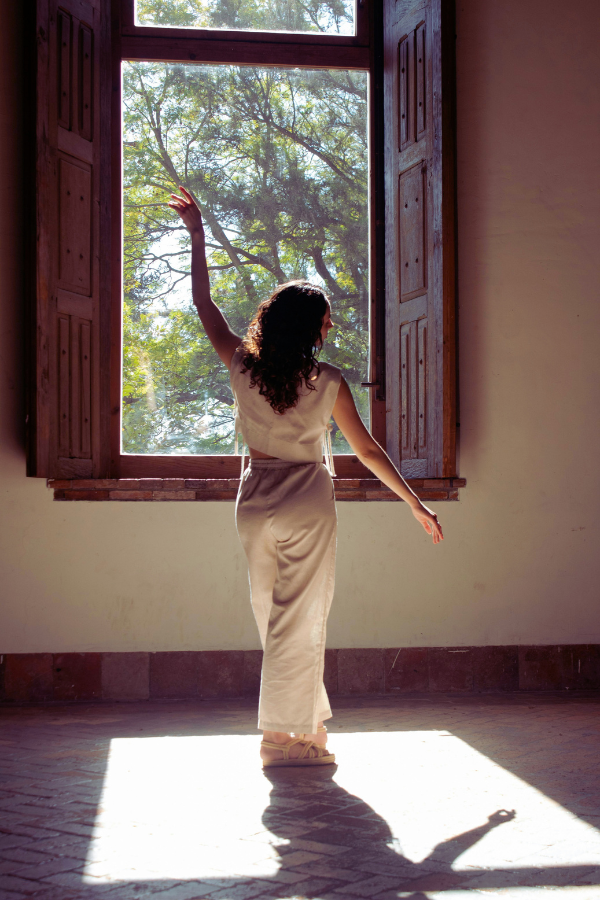 A person in light-colored clothing stands barefoot in a sunlit room, gently reaching one arm upward toward a large window with open shutters. Their shadow is cast on the floor.