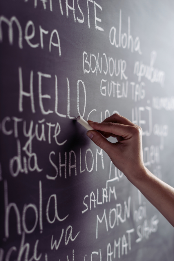 A hand writes the word “hello” in chalk on a blackboard surrounded by greetings in multiple languages, including “bonjour,” “hola,” and “shalom.”
